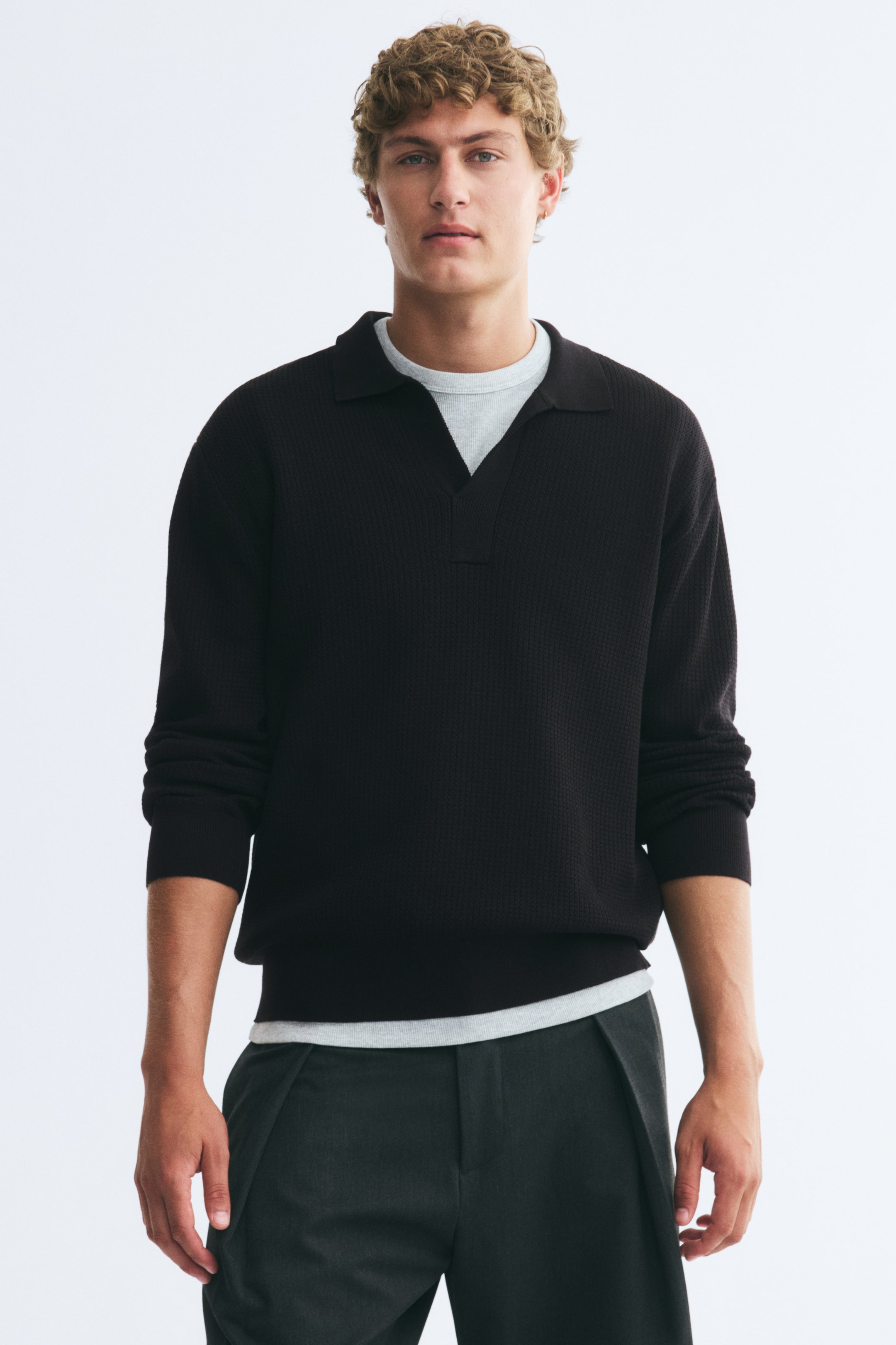 View larger image: A black textured-knit polo shirt with an open collar, long sleeves, and a ribbed hem is worn by the model. The relaxed-fit polo is layered over a light gray T-shirt, which is visible at the collar and hem, and styled with dark pleated trousers.
