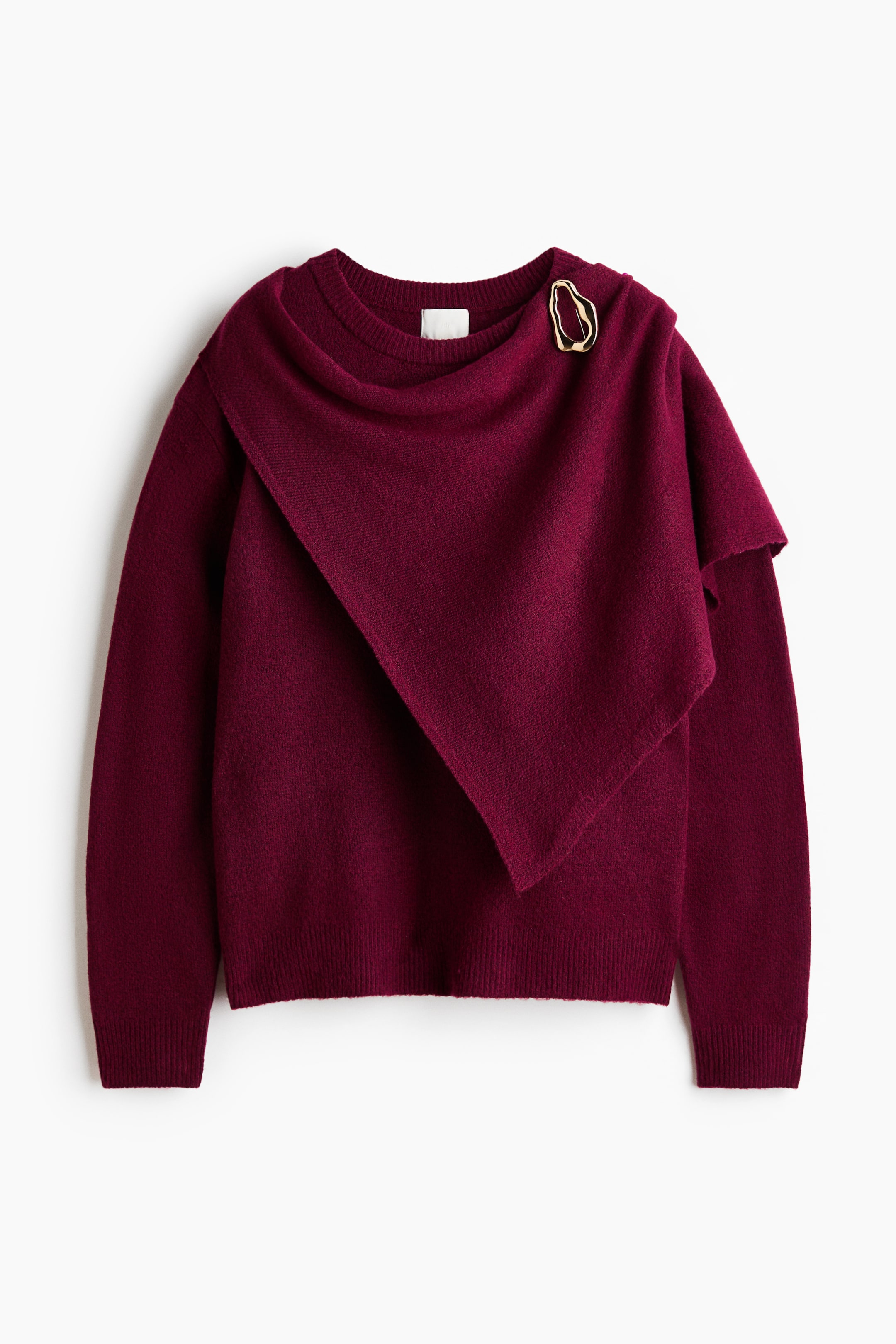 View larger image: A deep red knit sweater featuring an asymmetrical draped panel across the front, secured at the right shoulder with a gold-toned, irregular-shaped brooch. The sweater displays ribbed cuffs and a ribbed hem.