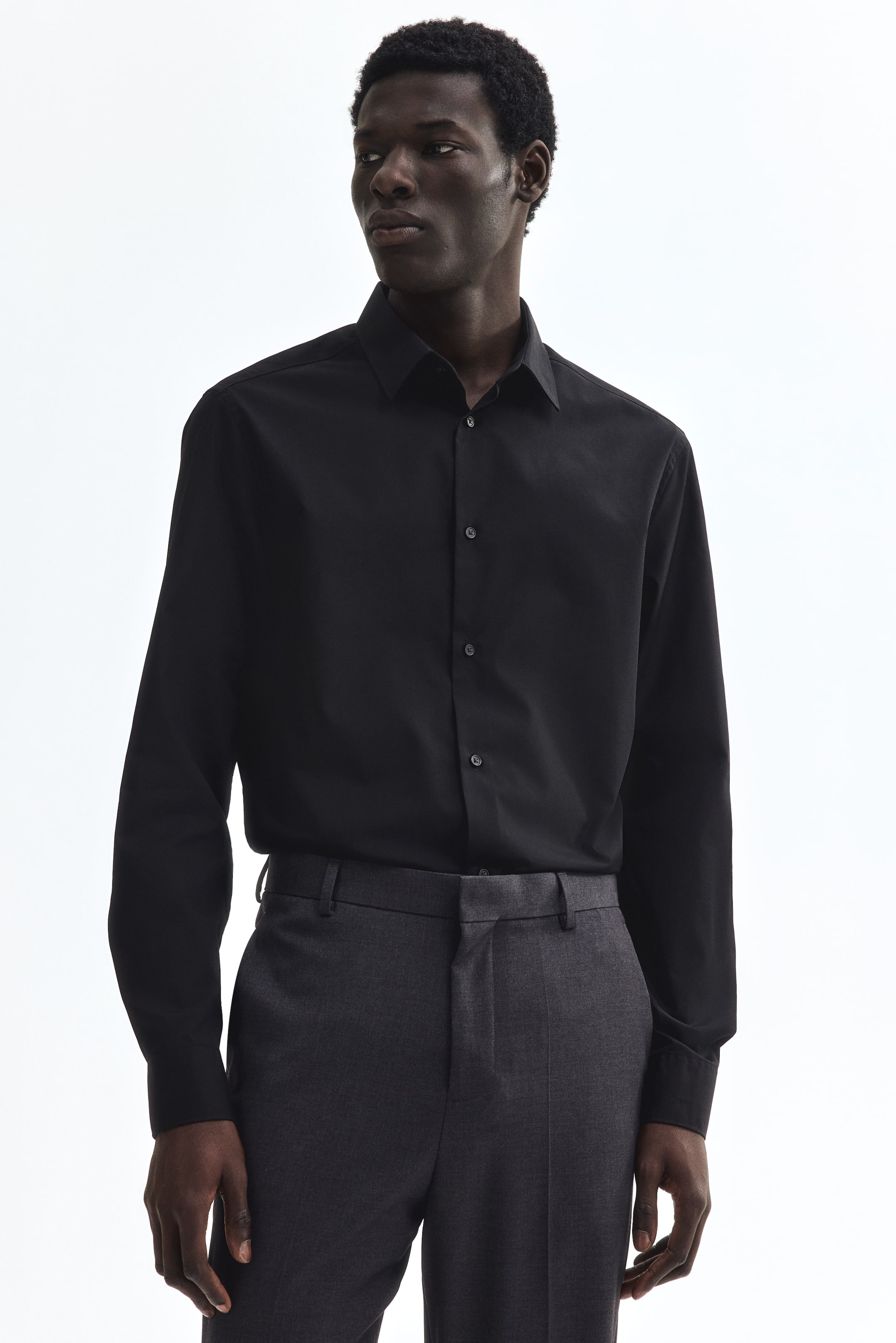 Voir une image plus grande: Une chemise noire à boutons présente un col évasé, des manches longues avec des poignets boutonnés et une patte de boutonnage à l'avant avec des boutons foncés. Elle est portée, rentrée dans un pantalon gris foncé, et met en valeur sa coupe ajustée sous un léger angle avant-droit.