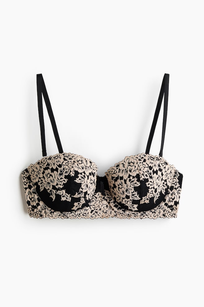 A strapless balconette bra is displayed from the front, showcasing black fabric with a beige floral lace overlay on the cups and band. Black, adjustable, detachable shoulder straps are extending upwards. - 1