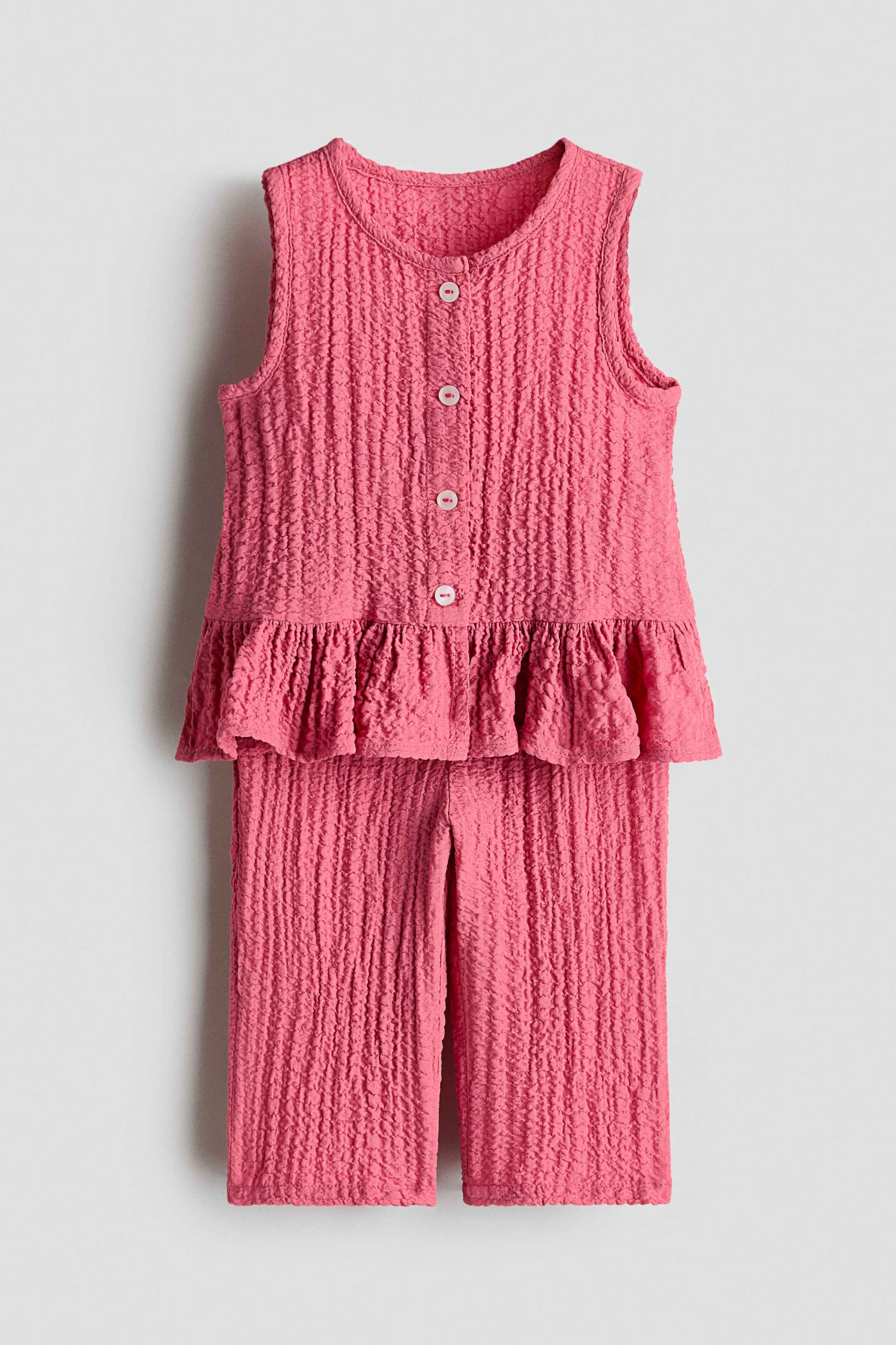 View larger image: A pink seersucker set, presented front-facing, displays a sleeveless top with a round neckline, five white buttons down the front and a wide frill at the hem. The matching trousers show a straight leg with a small frill trim at the elasticated waist.