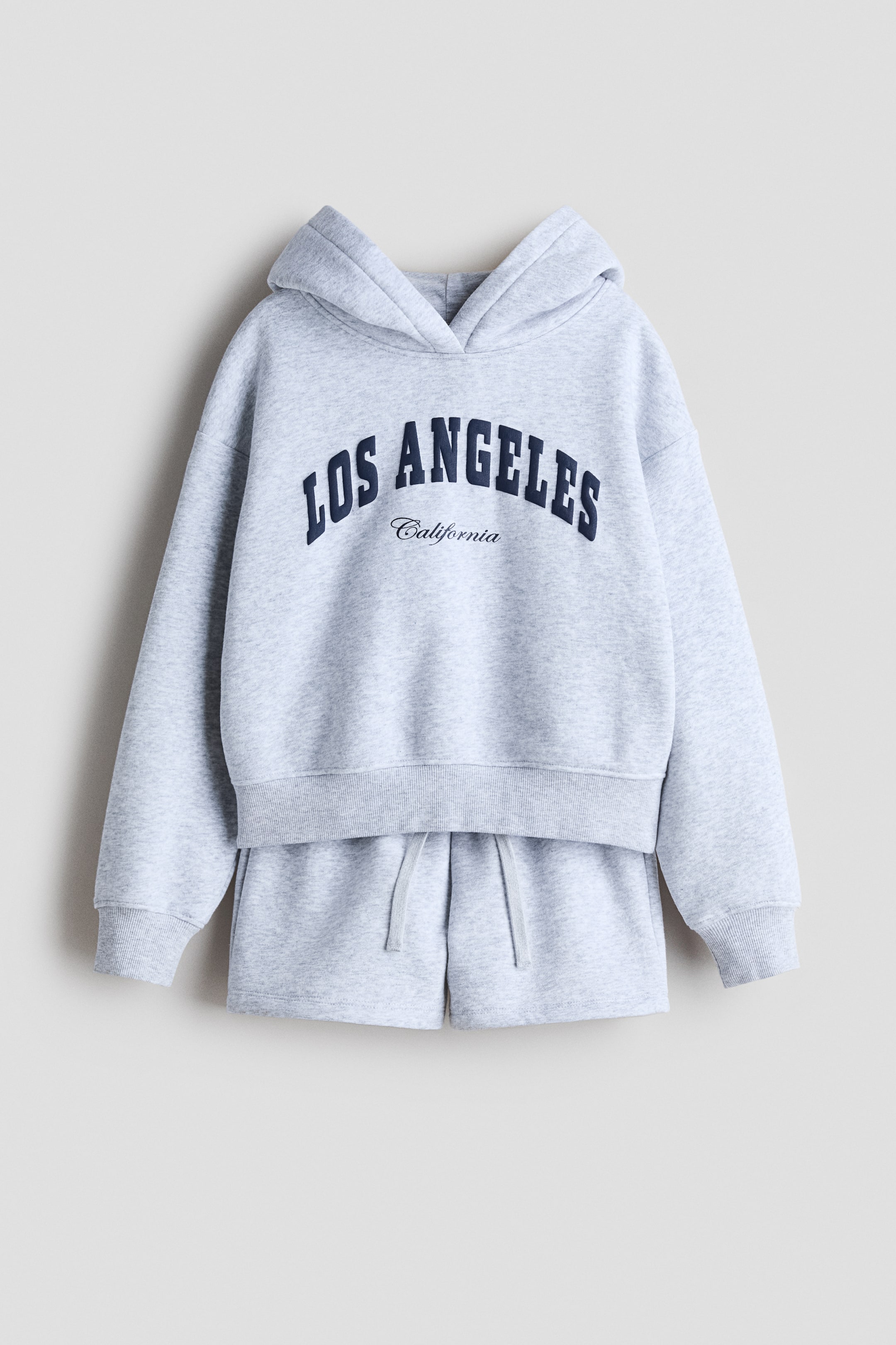 View larger image: A light grey sweatshirt fabric set is featuring a hoodie and matching shorts, both presented from the front. The hoodie is displaying a dark blue 'LOS ANGELES California' print motif on the chest, a lined hood with a wrapover front, ribbed cuffs, and a ribbed hem, while the shorts are designed with an elasticized, drawstring waist.