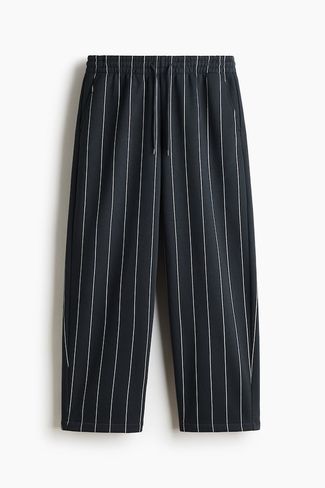 Track pants in a dark grey fabric with thin white vertical pinstripes are shown from a front-facing view. The pants feature a relaxed, straight-leg fit, an elasticated waistband with matching drawstrings, and diagonal side pockets. - 1