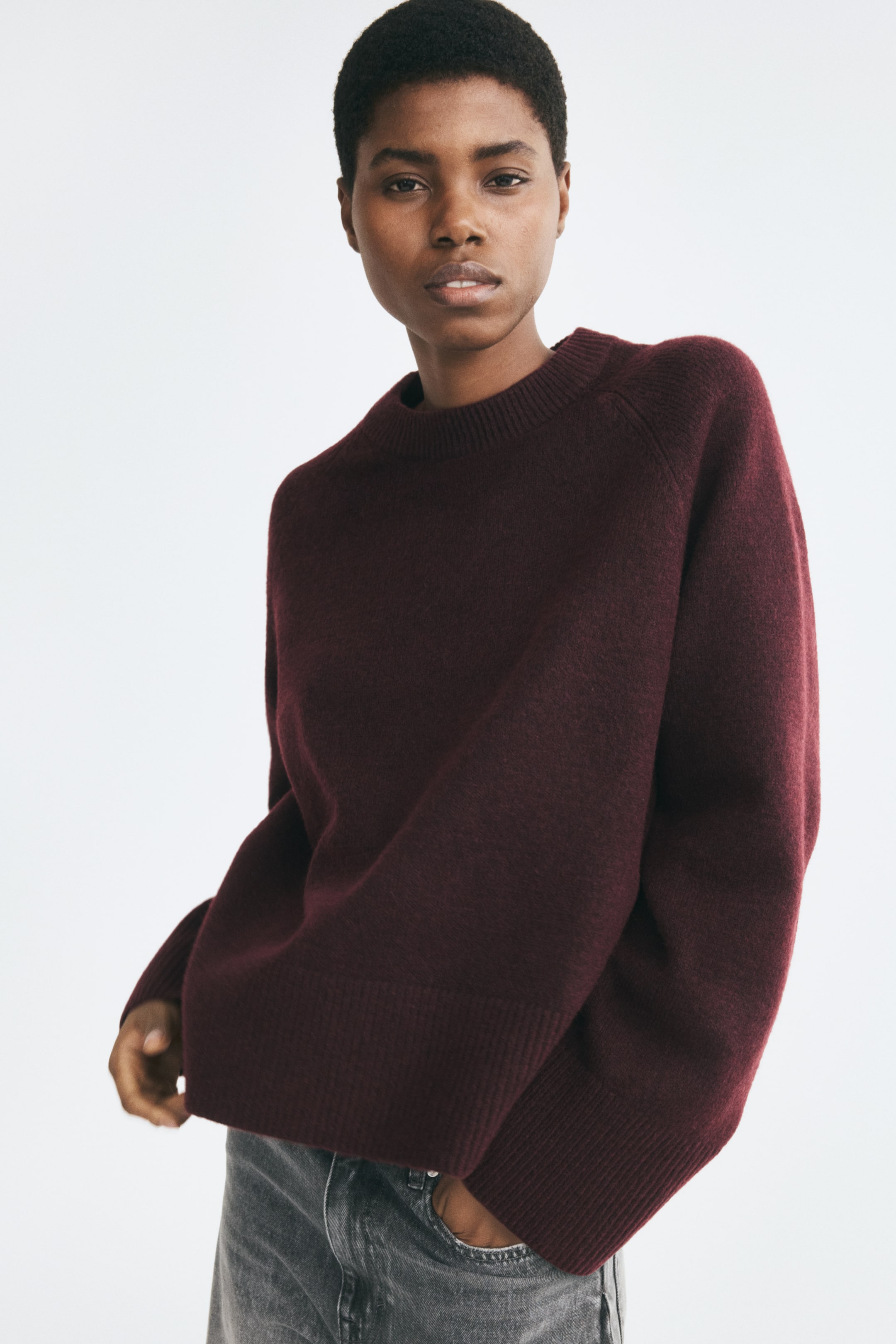 View larger image: The model is wearing a burgundy knit sweater featuring a round, rib-trimmed neckline, long raglan sleeves, and wide ribbed cuffs and hem. She is posing with one hand touching the hem of the sweater, styled with grey jeans.