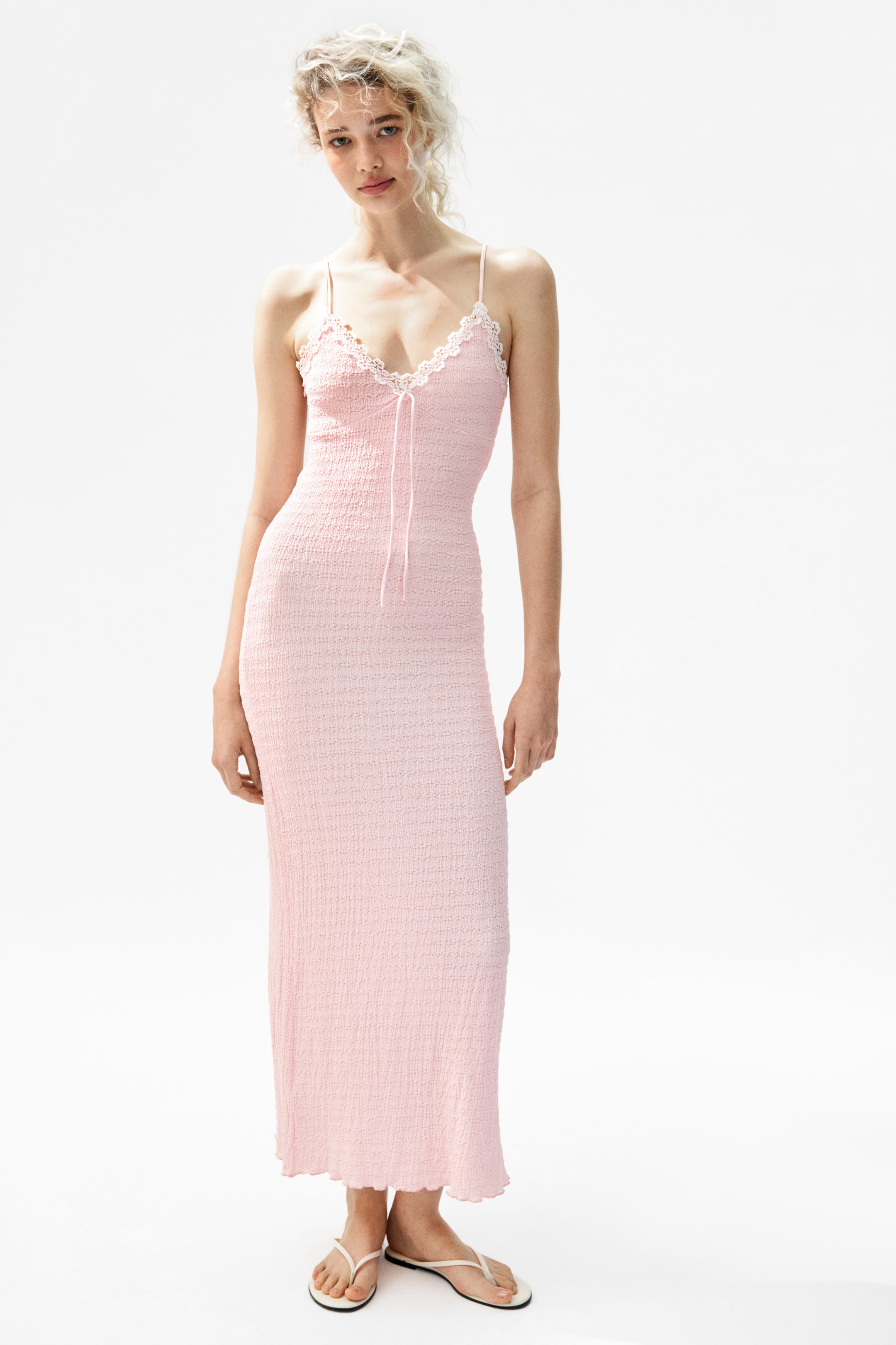 Größeres Bild ansehen: Ein rosa gesmoktes Midi-Schlupfkleid, das ein V-Ausschnitt mit weißer Spitzenverzierung, dünne Spaghettiträger, ein rosa Kordelzug in der Mitte des Busens und einen welligen Saum aufweist, wird von dem Model getragen.