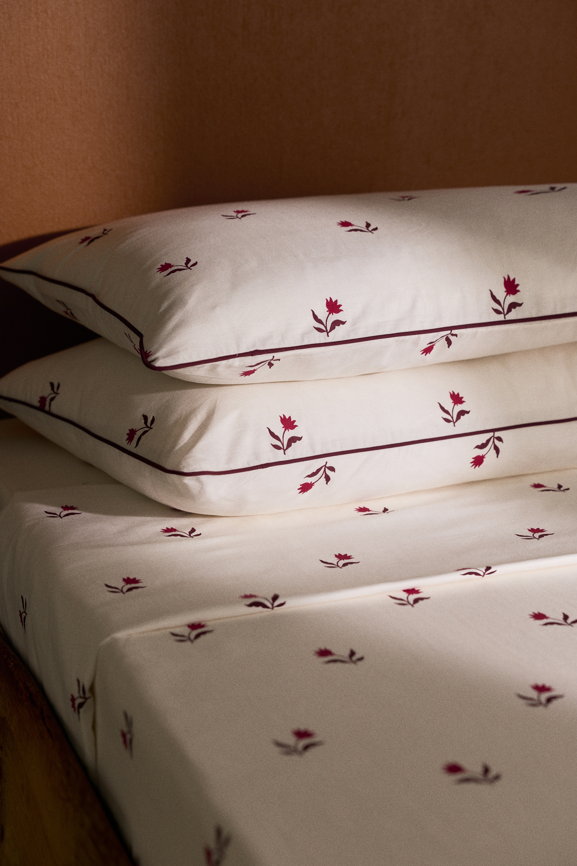 A white bedding set features a repeating pattern of small, dark red tulip motifs across the duvet cover and two matching pillowcases. The pillowcases feature thin, dark red piped borders along the edges and are presented from an overhead angle.