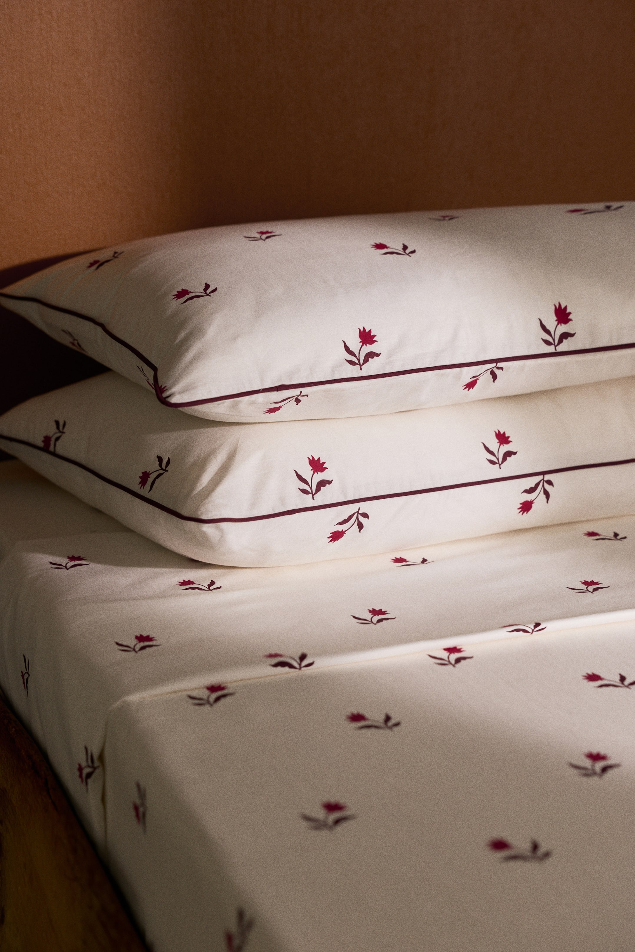 View larger image: A cotton quilt cover set features an off-white fabric with an all-over small red floral print and contrasting burgundy piping along the edges. The set includes two matching pillowcases stacked on top of the neatly laid quilt cover.