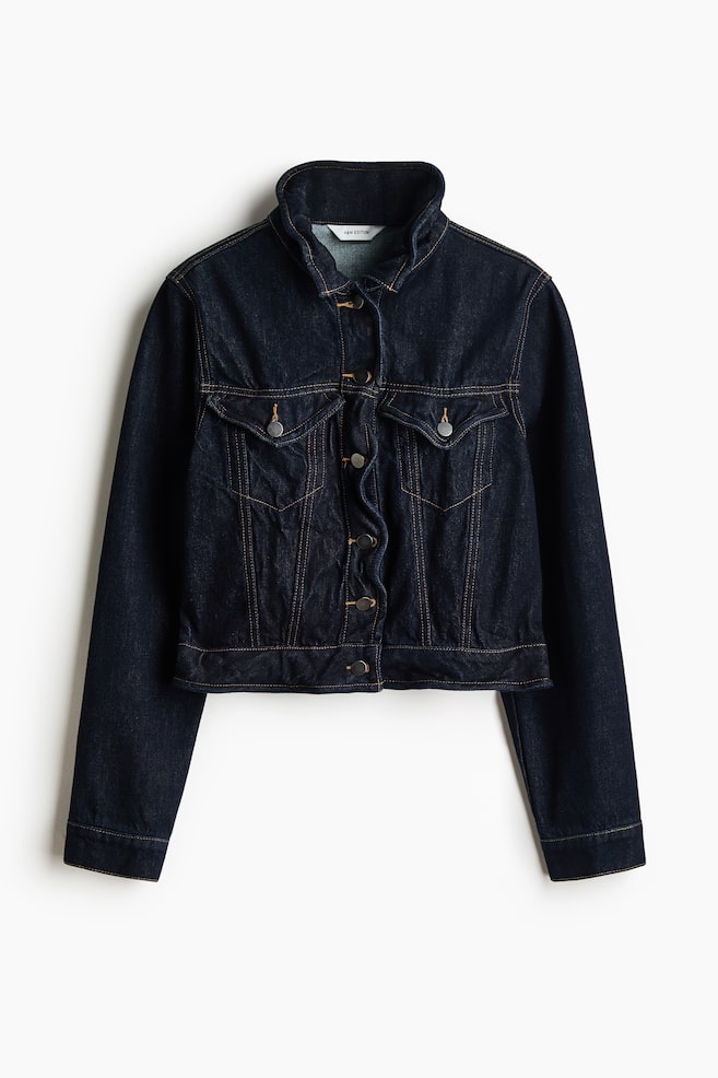 A short denim jacket in dark indigo blue is shown from the front, featuring a crumpled, structured collar and a wavy front button placket. The jacket has orange contrast stitching, two buttoned chest flap pockets, and a cropped waist. - 1