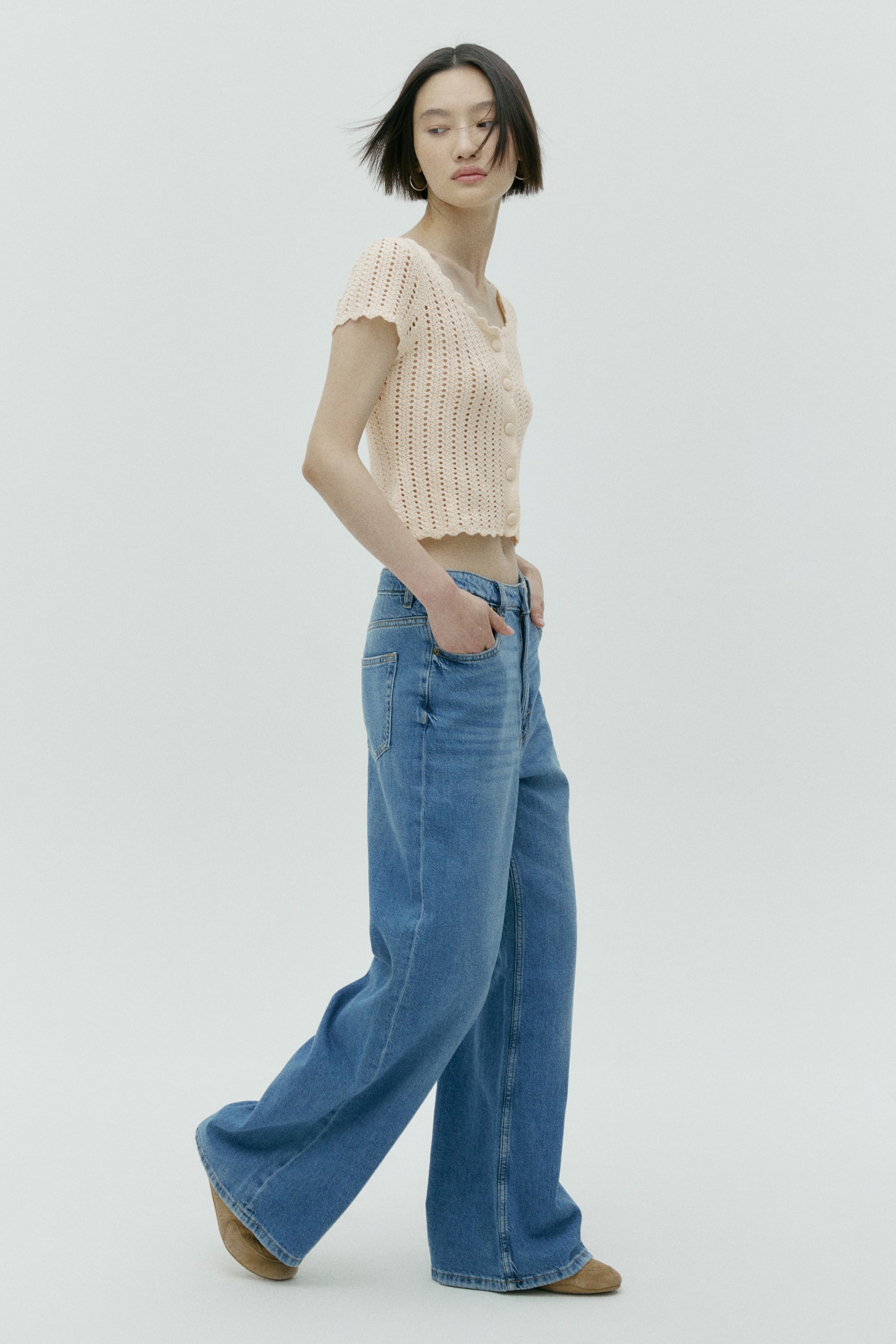 View larger image: A light peach cropped open-knit top displays short sleeves and a button-front closure, featuring scalloped edges along its neckline, sleeves, and hem. It is styled with high-waisted, wide-leg blue denim jeans that show a relaxed fit from a slightly front-side angle.
