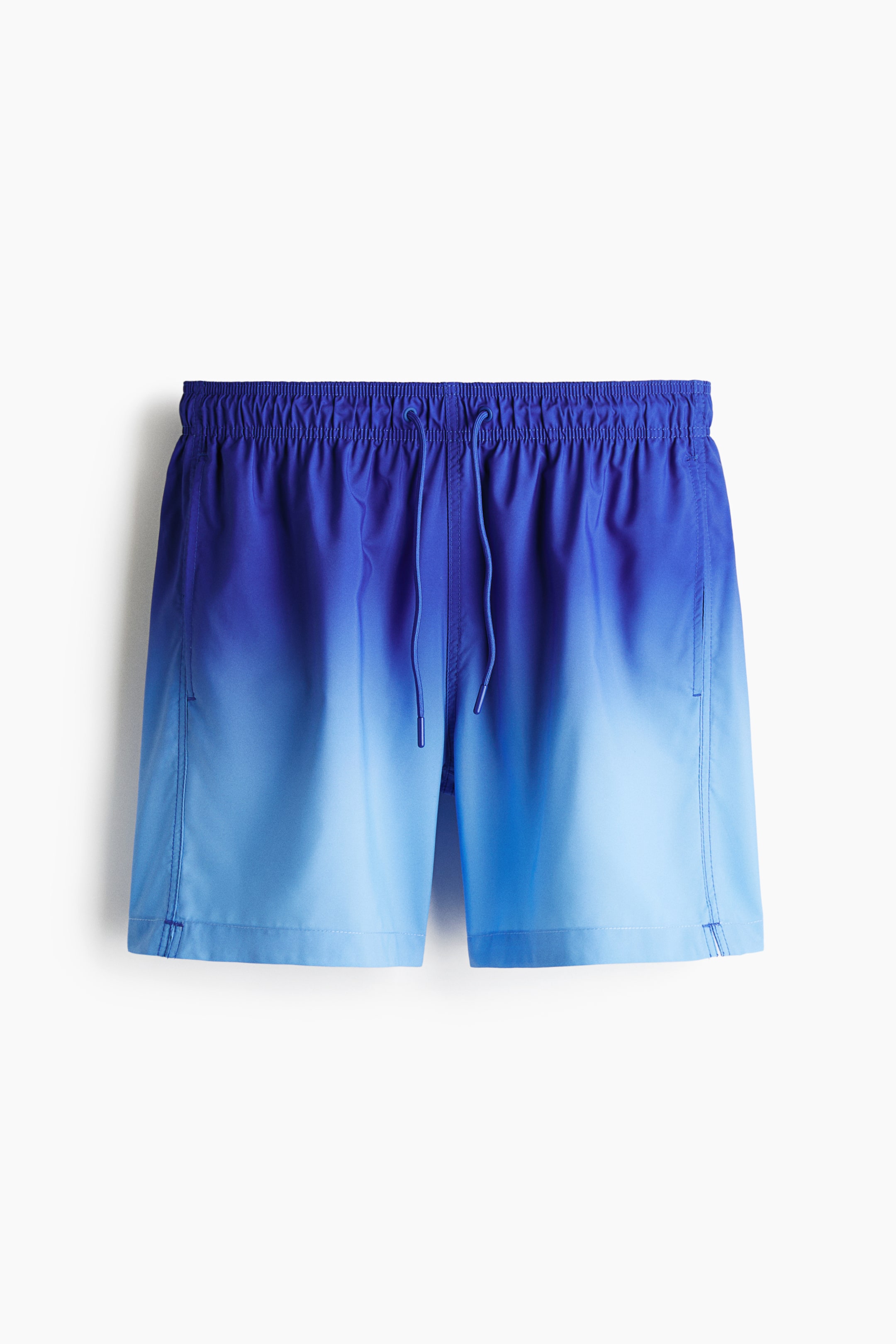 View larger image: Swim shorts are facing FRONT, showcasing an ombre pattern that transitions from a deep blue at the waist to a lighter blue at the hem. The waist is designed with covered elastication and a visible drawstring, while side pockets are included, featuring a small slit detail on one side of the hems.
