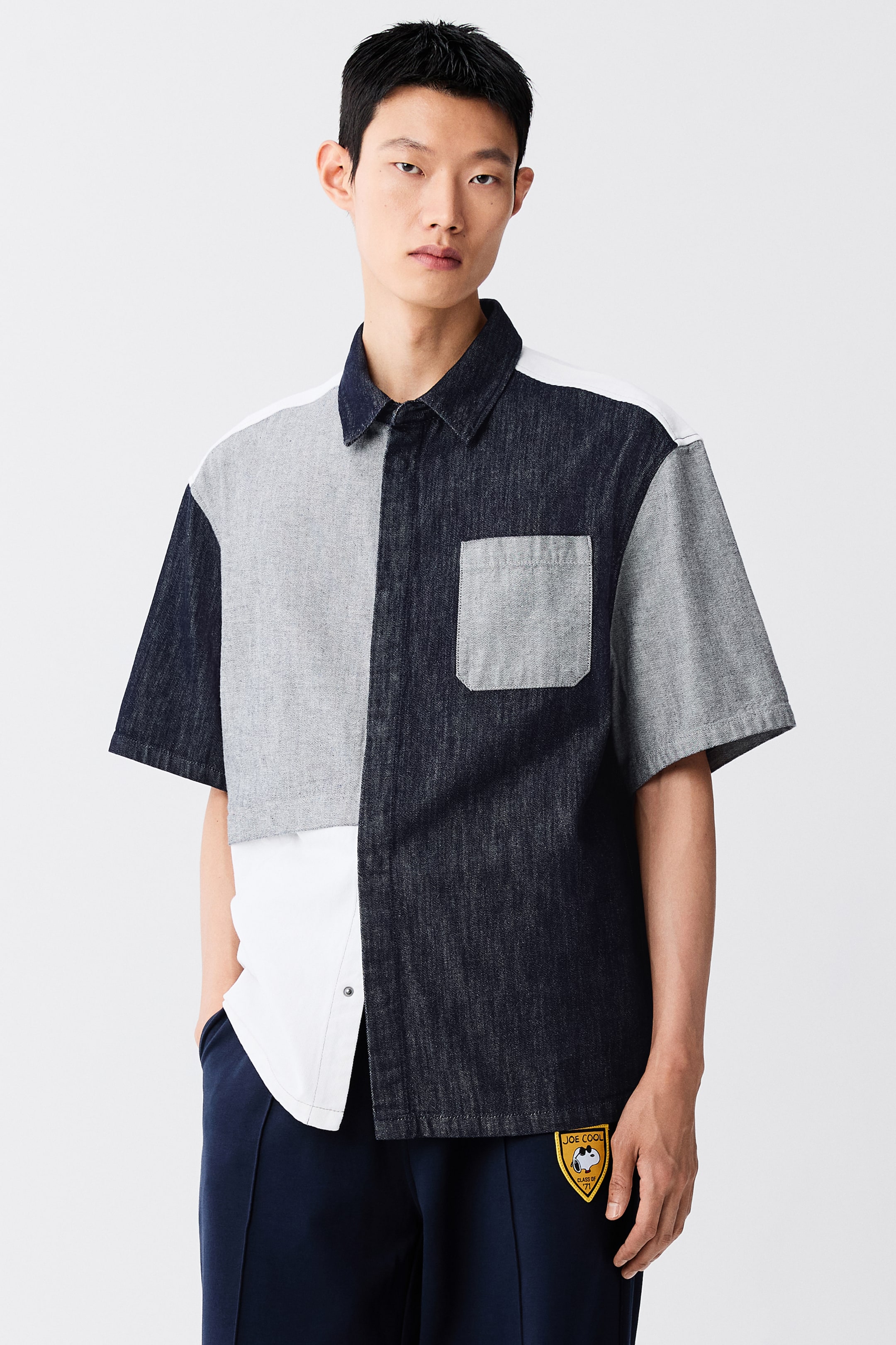 View larger image: The model is wearing a short-sleeve, colour-block denim shirt in dark blue, light grey, and white panels. The loose-fit shirt features a classic collar and a light grey patch pocket on the chest, paired with dark blue trousers.