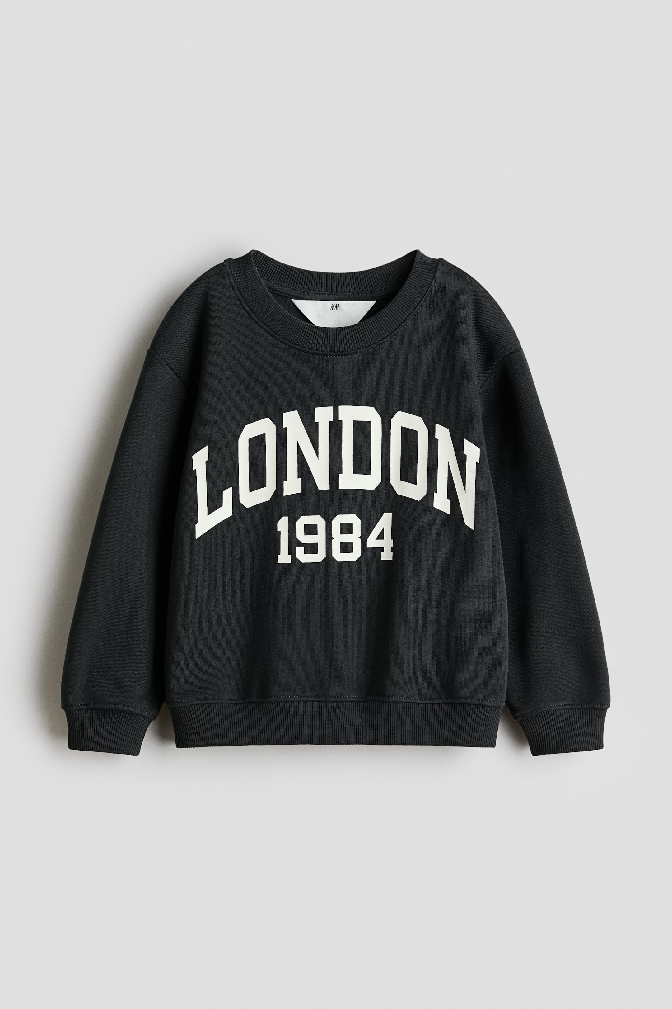 View larger image: A dark gray sweatshirt is lying flat, presenting its front view and featuring a white graphic print on the chest. The print is displaying 'LONDON' in large arched block letters above '1984', and the sweatshirt is designed with a crew neck, ribbed cuffs, and a ribbed hem.