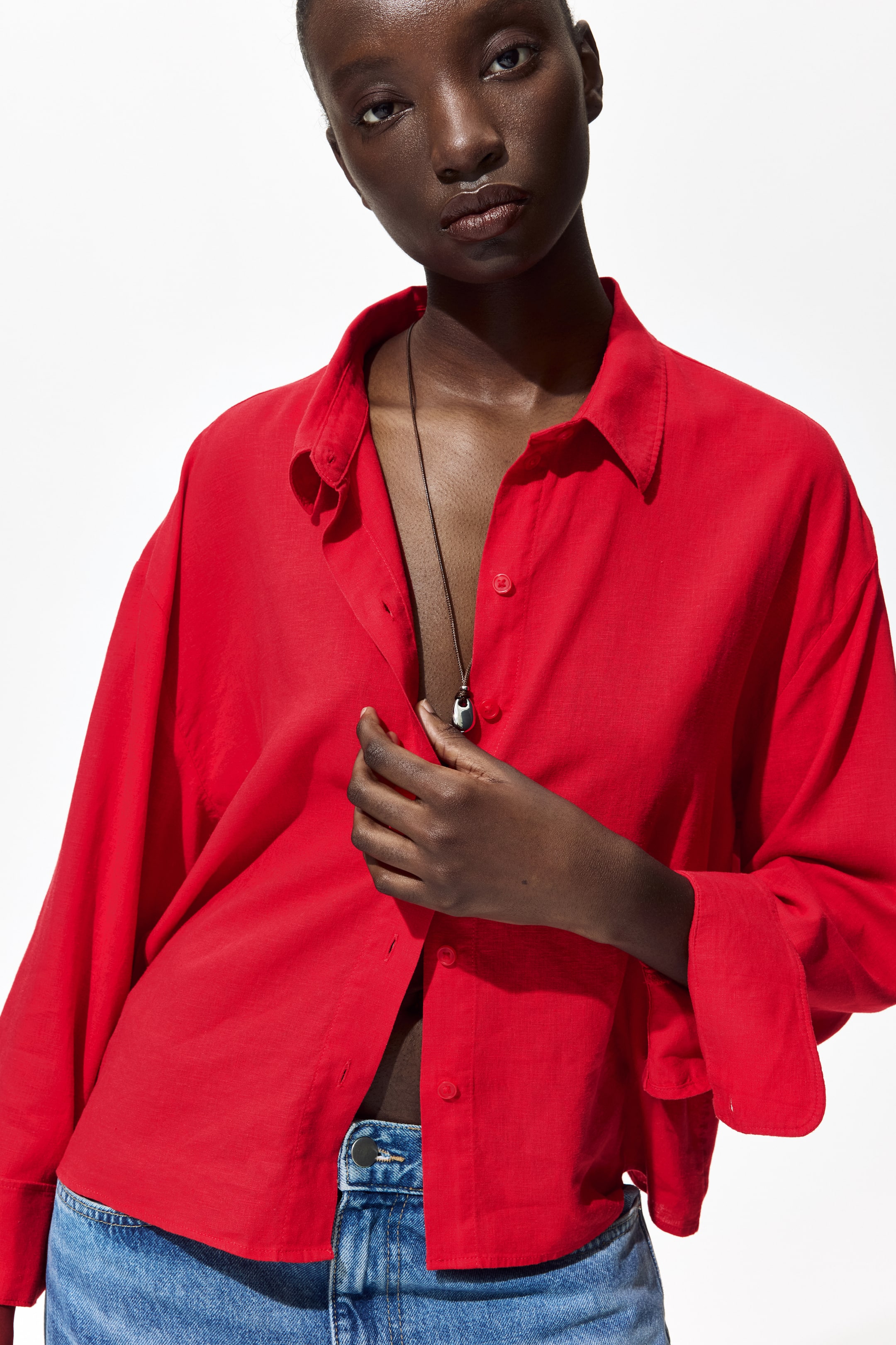 View larger image: A red button-up shirt features a pointed collar and long, relaxed sleeves. Its front placket is slightly unbuttoned, showcasing a silver pendant necklace. The shirt is cropped, sitting just above blue denim jeans.