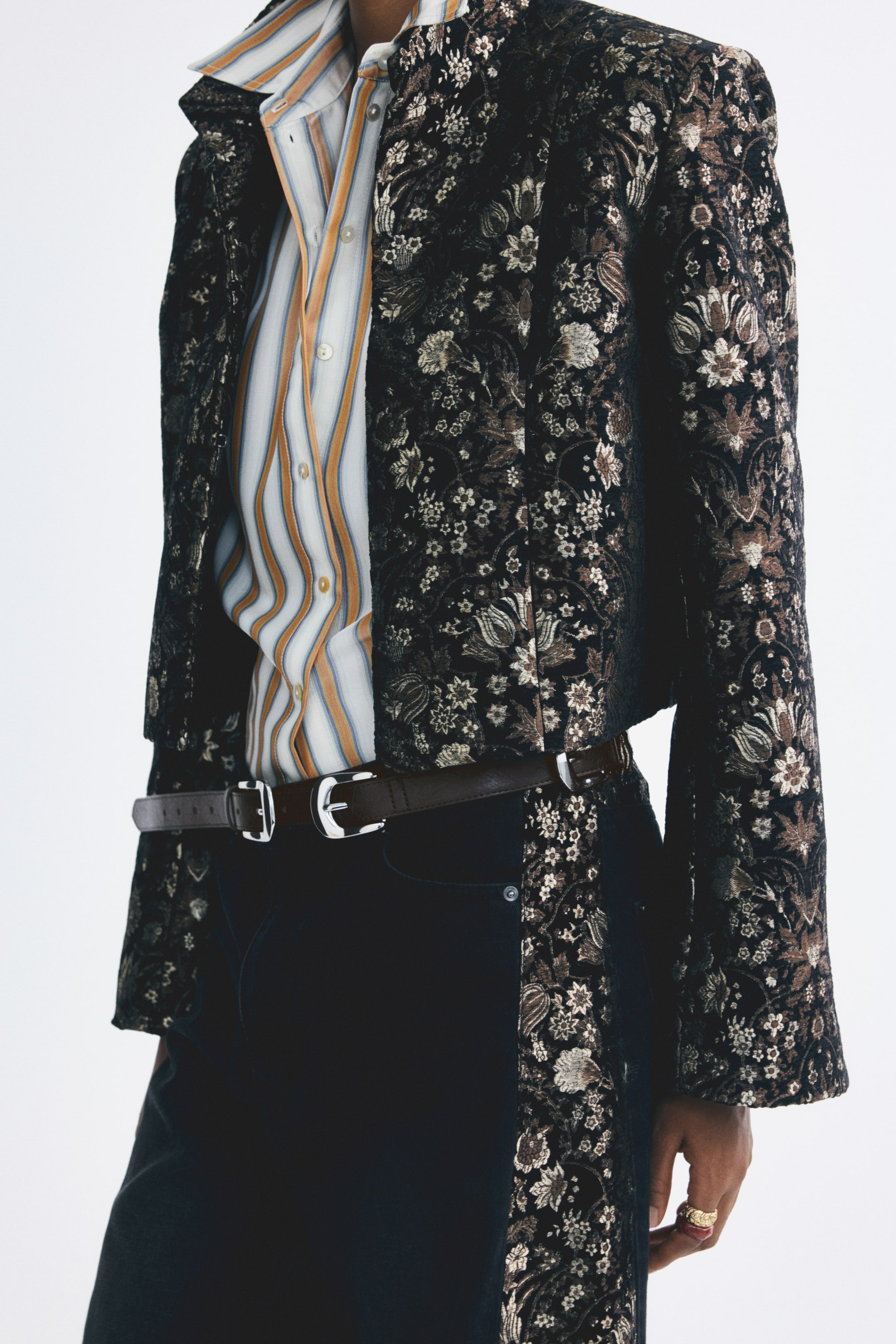 Agrandir l'image: Une ceinture marron foncé en tissu enduit, ornée de plusieurs boucles en métal ton argent régulièrement espacées, est portée dans les passants d’un pantalon foncé par le mannequin. La ceinture est associée à une chemise rayée rentrée dans le pantalon et à une veste ouverte à motif floral.