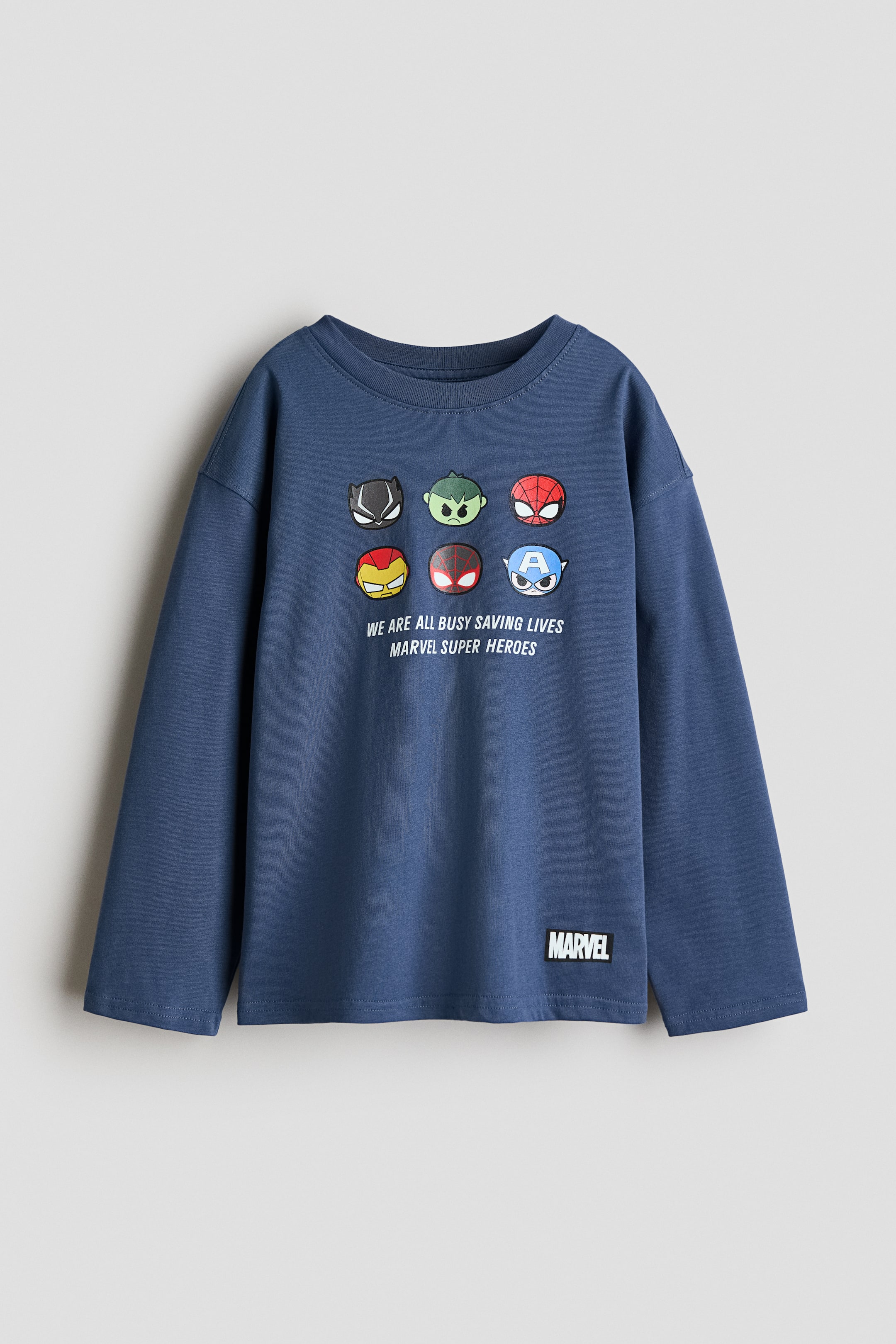 Grotere afbeelding bekijken: Een blauwe T-shirt met lange mouwen is van voren afgebeeld, met een ronde hals met ribboord en een grafische print van zes cartoon-superheldengezichten op de borst. De print toont de tekst "WE ARE ALL BUSY SAVING LIVES MARVEL SUPER HEROES" in wit, met een klein zwart merklogo-label dat dicht bij de onderzoom is vastgenaaid.