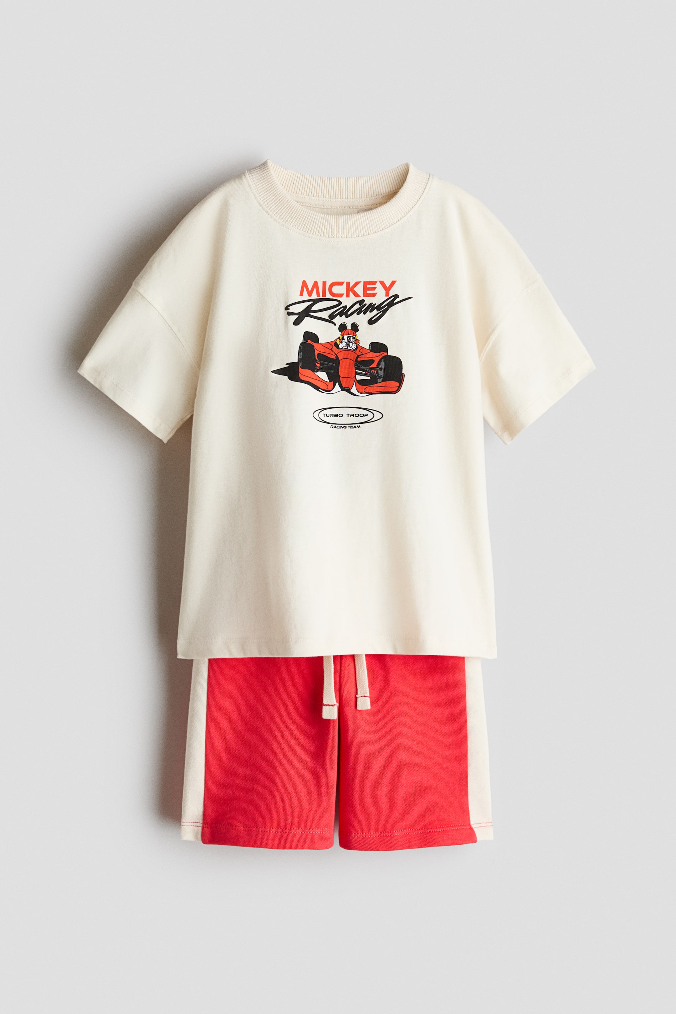 Grotere afbeelding bekijken: Een tweedelig kindersetje toont een off-white T-shirt en rode shorts, beide aan de voorkant. Het T-shirt heeft een geribde ronde hals, korte mouwen en een "Mickey Racing"-afbeelding met Mickey Mouse in een rode raceauto en de tekst "Turbo Troop Racing Team", terwijl de shorts rode stof met off-white zijstrepen en een zichtbare off-white trekkoord bij de elastische taille hebben.