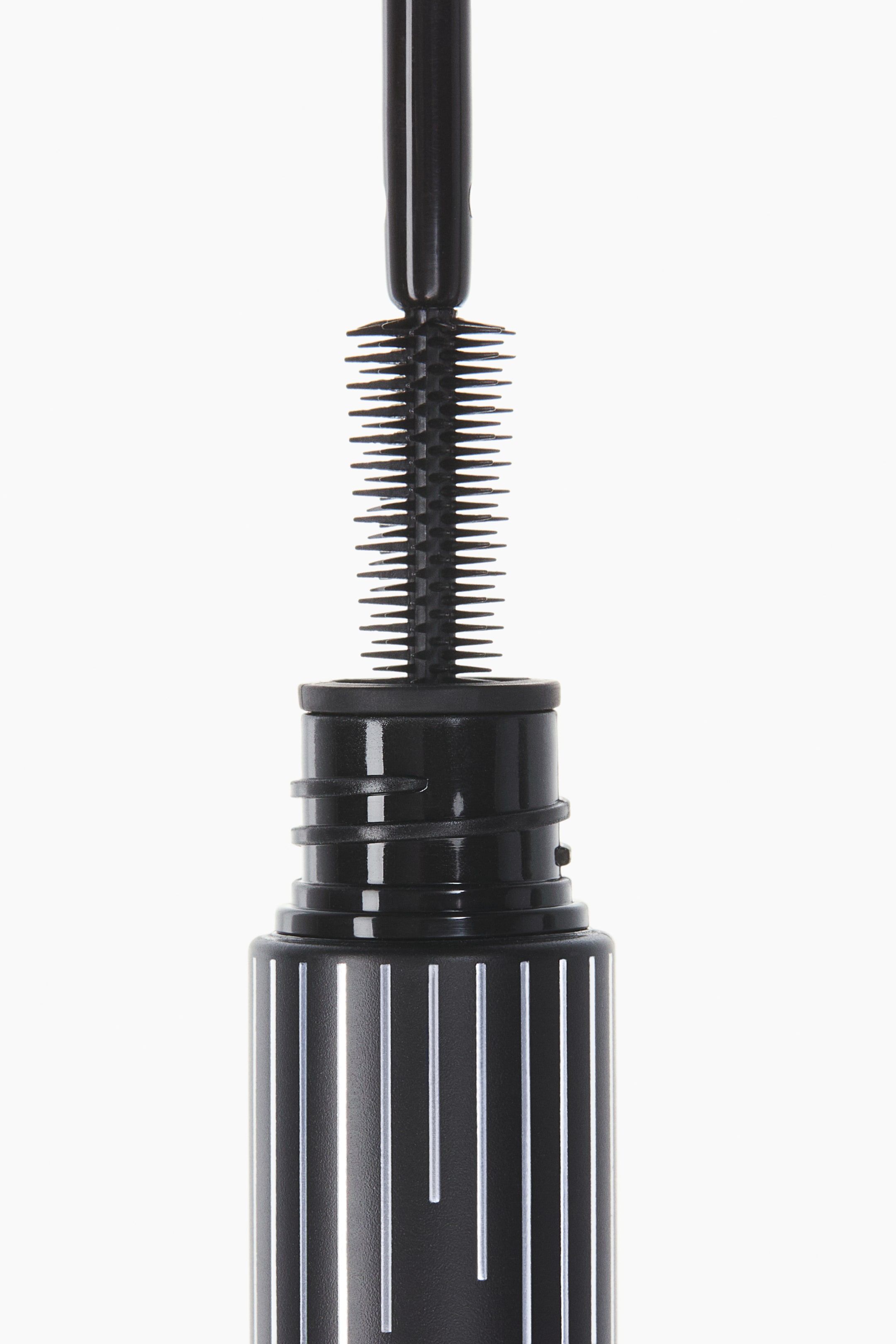 View larger image: A black mascara wand featuring a brush with short, pointed silicone bristles is extending from its black container. A black wiper mechanism is visible at the container's neck, and its body is patterned with vertical white stripes of varying lengths.