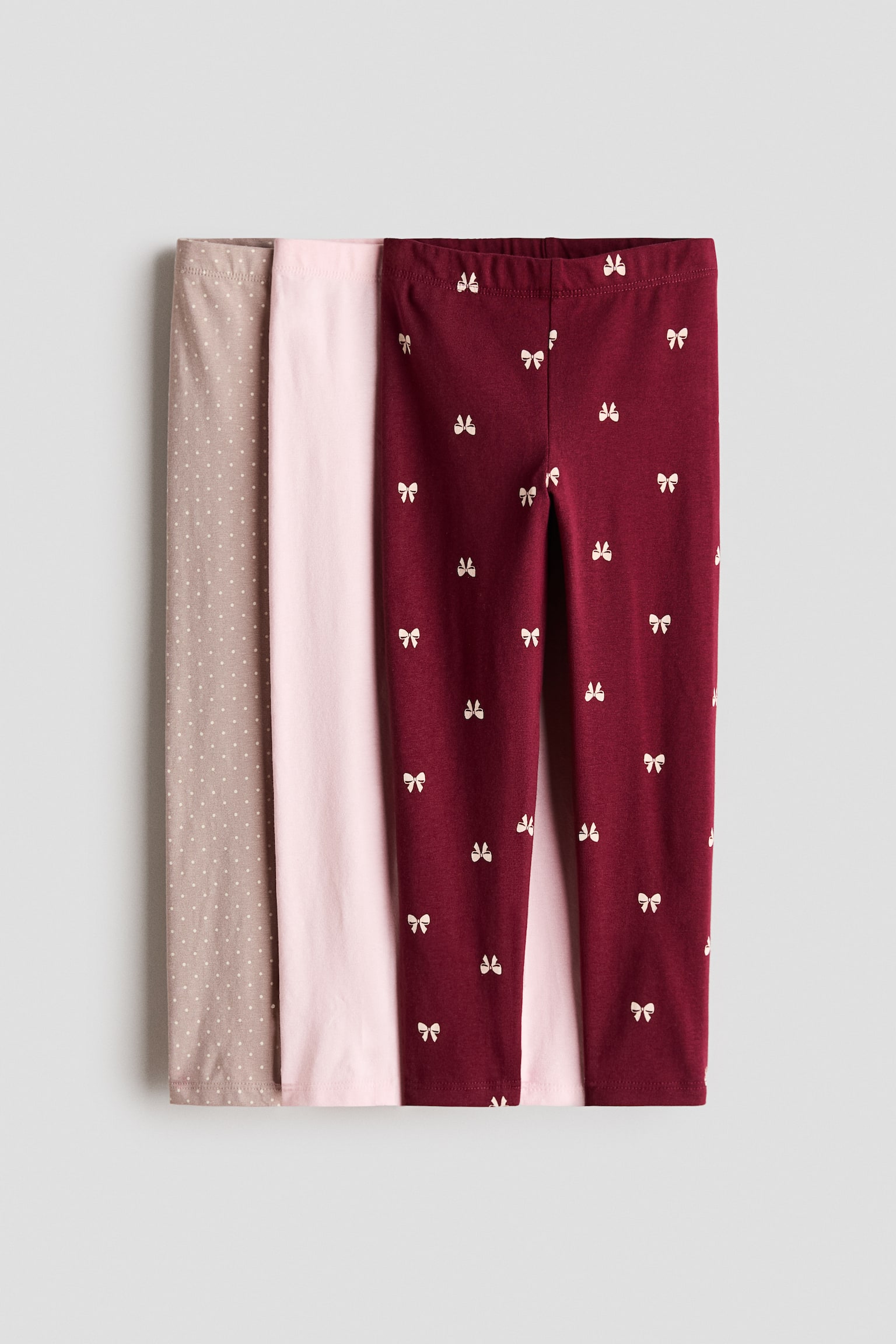 Ein Dreierpack Leggings aus Baumwolljersey wird von vorne prÃ¤sentiert und verfÃ¼gt Ã¼ber einen elastischen Bund sowie schmale Beine. Das vorderste Paar ist dunkelrot und mit einem wiederholenden Muster aus kleinen weiÃen Schleifen versehen, wÃ¤hrend dahinter ein einfarbiges hellrosa Paar und ein taupefarbenes Paar mit kleinen weiÃen Punkten Ã¼bereinandergelegt sind.