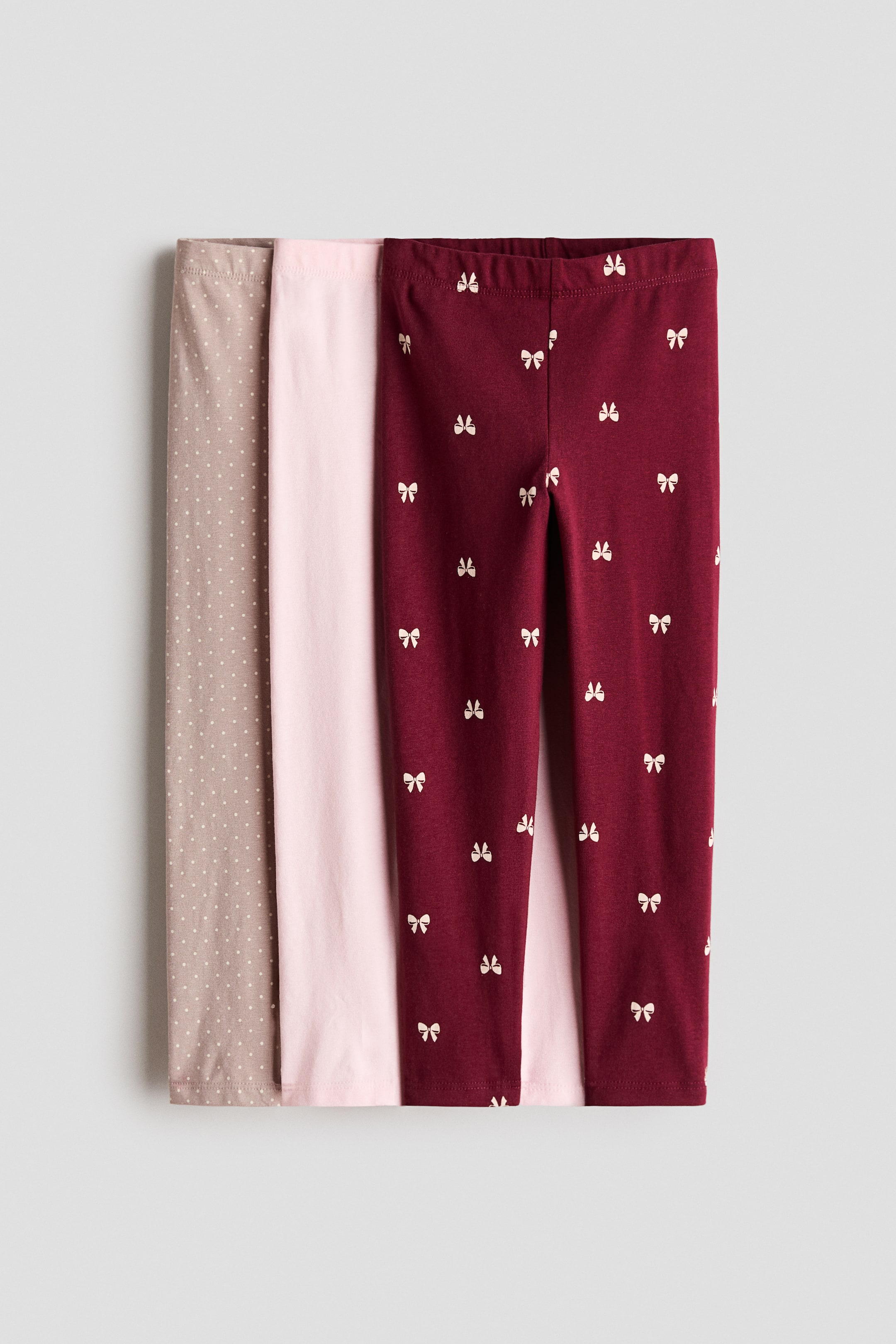 Agrandir l'image: Un lot de trois leggings en jersey de coton est présenté de face, avec une taille élastiquée et des jambes slim. La paire au premier plan est rouge foncé avec un motif répétitif de petits nœuds blancs, tandis qu’une paire rose clair unie et une paire taupe à petits pois blancs sont superposées derrière.