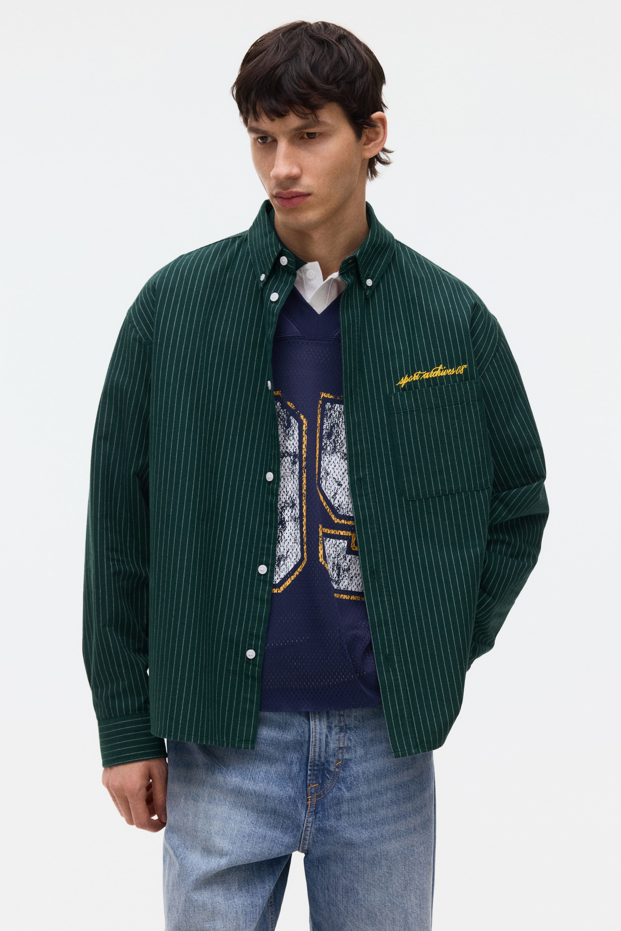 View larger image: A dark green pinstripe shirt, featuring a button-down collar, white button-front closure, and a left chest patch pocket with yellow embroidered text, is being worn open over a layered navy top, displaying its relaxed fit from a front-facing angle.