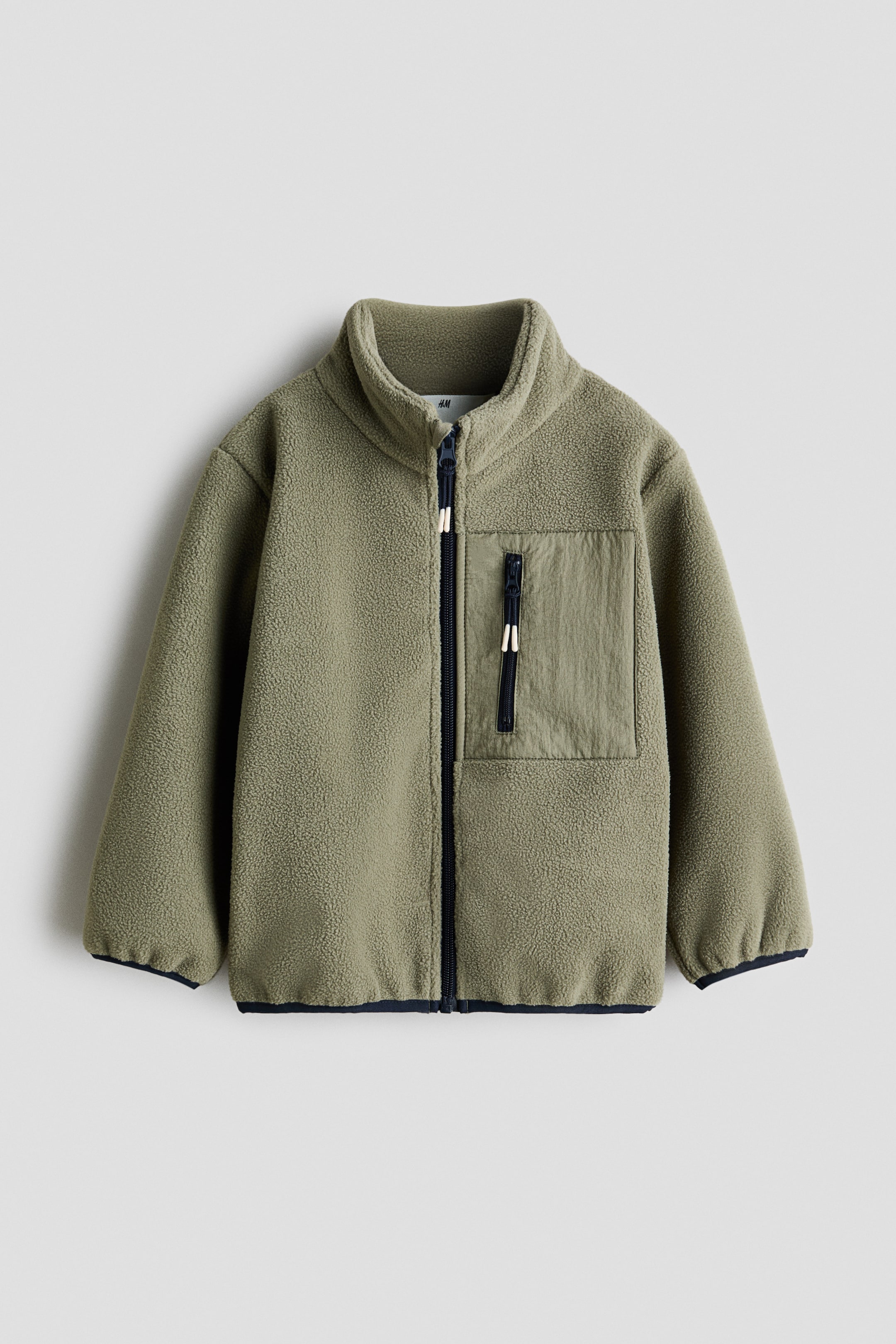 Größeres Bild anzeigen: Eine salbeigrüne Fleecejacke wird von vorne gezeigt, mit einem Stehkragen und einem durchgehenden Frontreißverschluss mit dunklem Band. Auf der linken Seite des Trägers ist eine kontrastierende Brusttasche aus Stoff mit einem vertikalen dunklen Reißverschluss und weißen Zippern zu sehen, und dunkle Einfassungen säumen die Bündchen und den Saum.