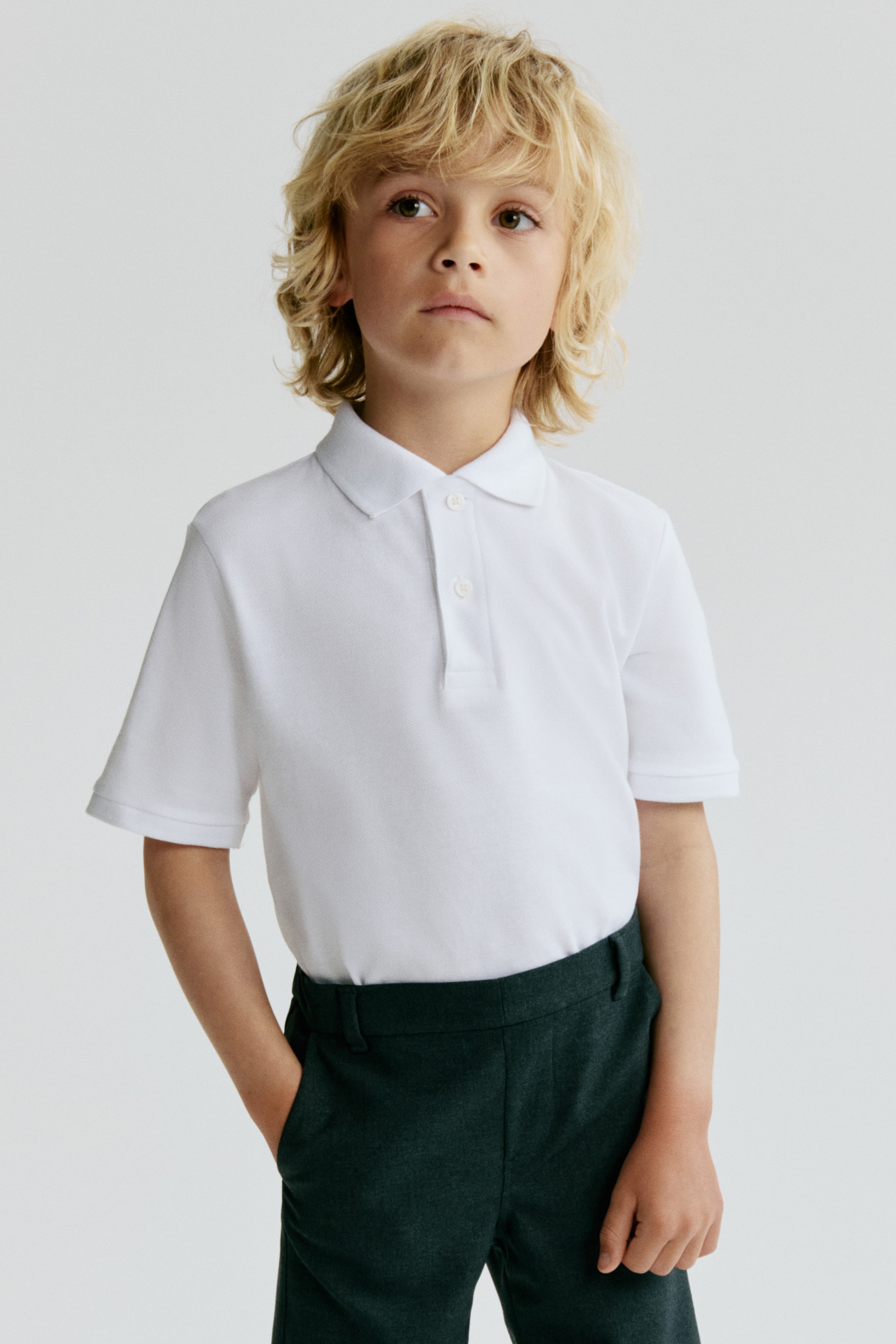 View larger image: A white polo shirt features a classic polo collar and a two-button placket with white buttons. It has short sleeves with ribbed cuffs and a textured pique knit fabric, presented tucked into dark trousers.