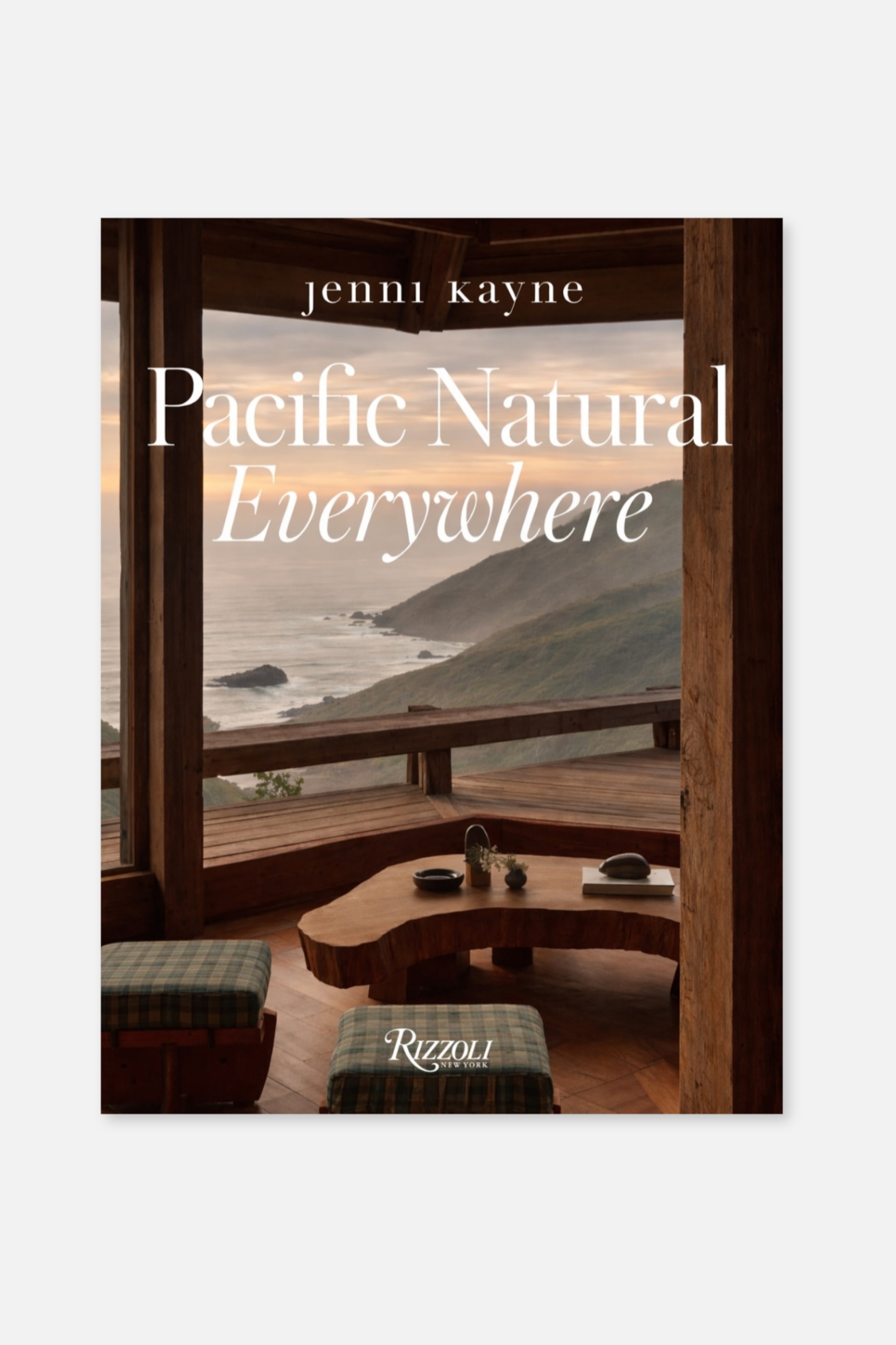 Größeres Bild anzeigen: Auf dem Buchcover wird der Titel "Pacific Natural Everywhere" in weißer Schrift dargestellt, mit "jenni kayne" darüber und "Rizzoli New York" darunter. Das Titelbild zeigt einen rustikalen Innenraum aus Holz, mit einem Tisch aus Holz mit natürlicher Kante und zwei karierten Ottomans, die auf eine malerische Küstenlinie und Berge blicken.