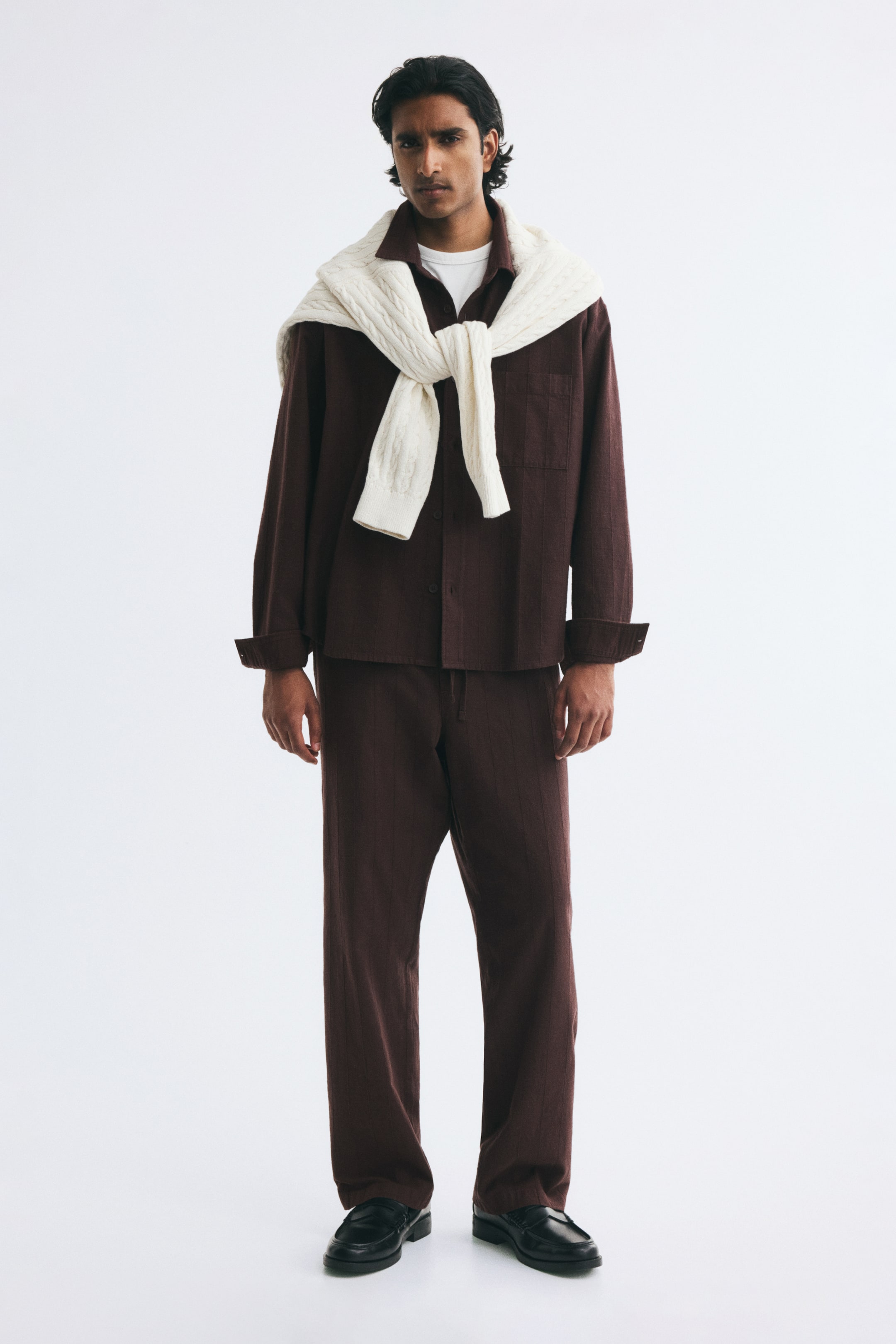 View larger image: The model is wearing relaxed-fit trousers in a dark brown textured cotton weave. The trousers are styled with a matching dark brown long-sleeve shirt, a white T-shirt, and a cream cable-knit sweater draped over the shoulders.