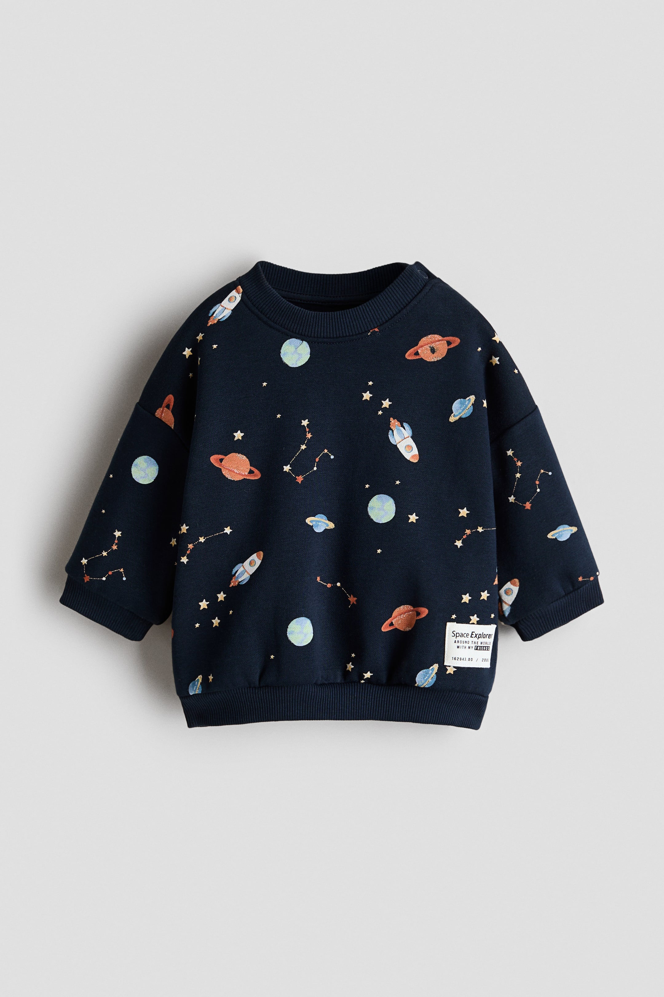 Voir une image plus grande: Chandail imprimé. Le chandail bleu marine présente un motif sur le thème de l'espace avec des fusées, des planètes, des étoiles et des constellations. Il est conçu avec un col rond, des manches longues, des poignets côtelés, un ourlet côtelé, et un écusson "Space Explorer" sur le bas à droite, présenté de face.