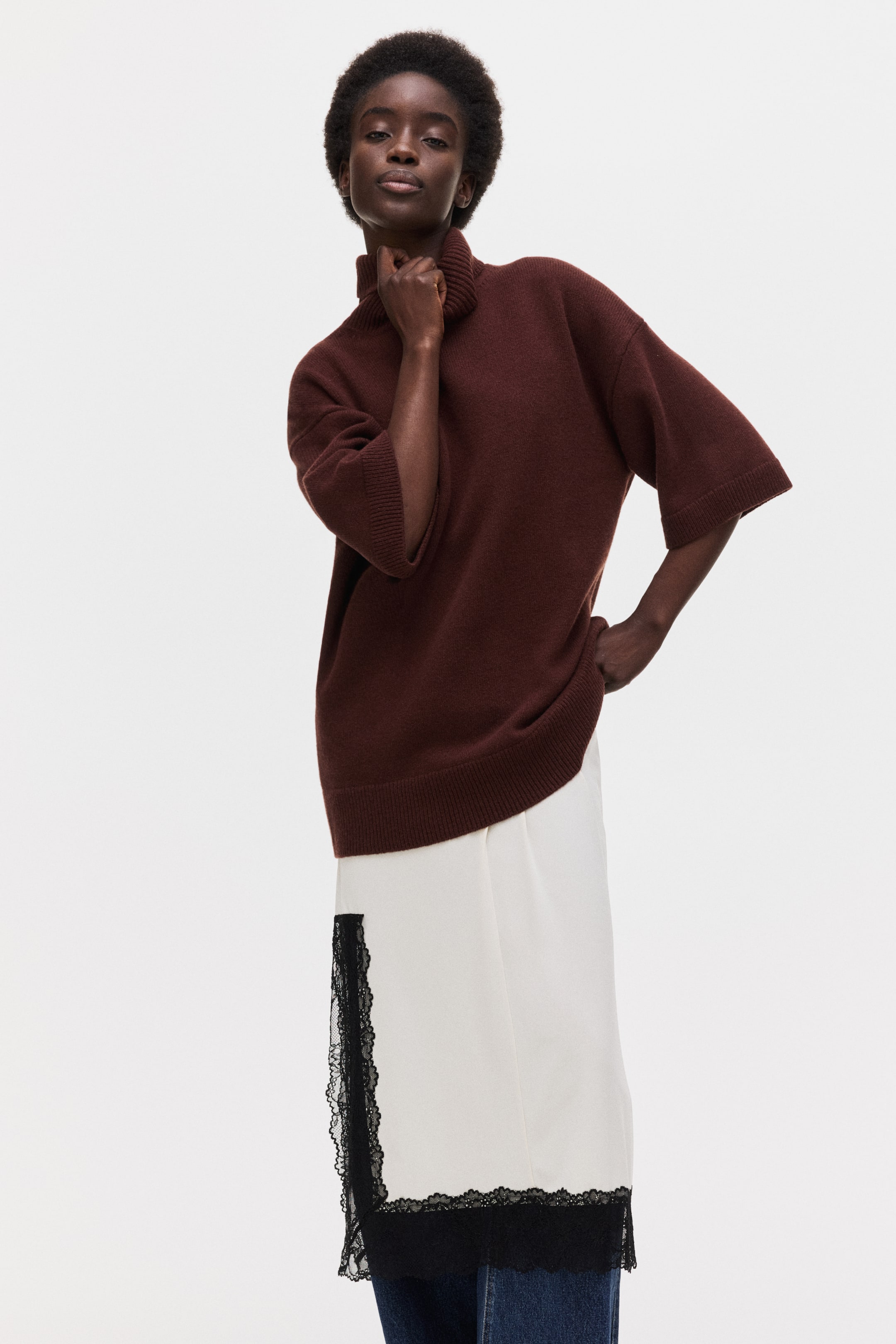 拡大画像を表示: ブラウンのタートルネックセーターは、ゆったりとしたフィット感、肘丈の袖にリブカフス、リブの裾を特徴としています。白いインナーの上に重ねられており、黒いレースのトリムが施されています。