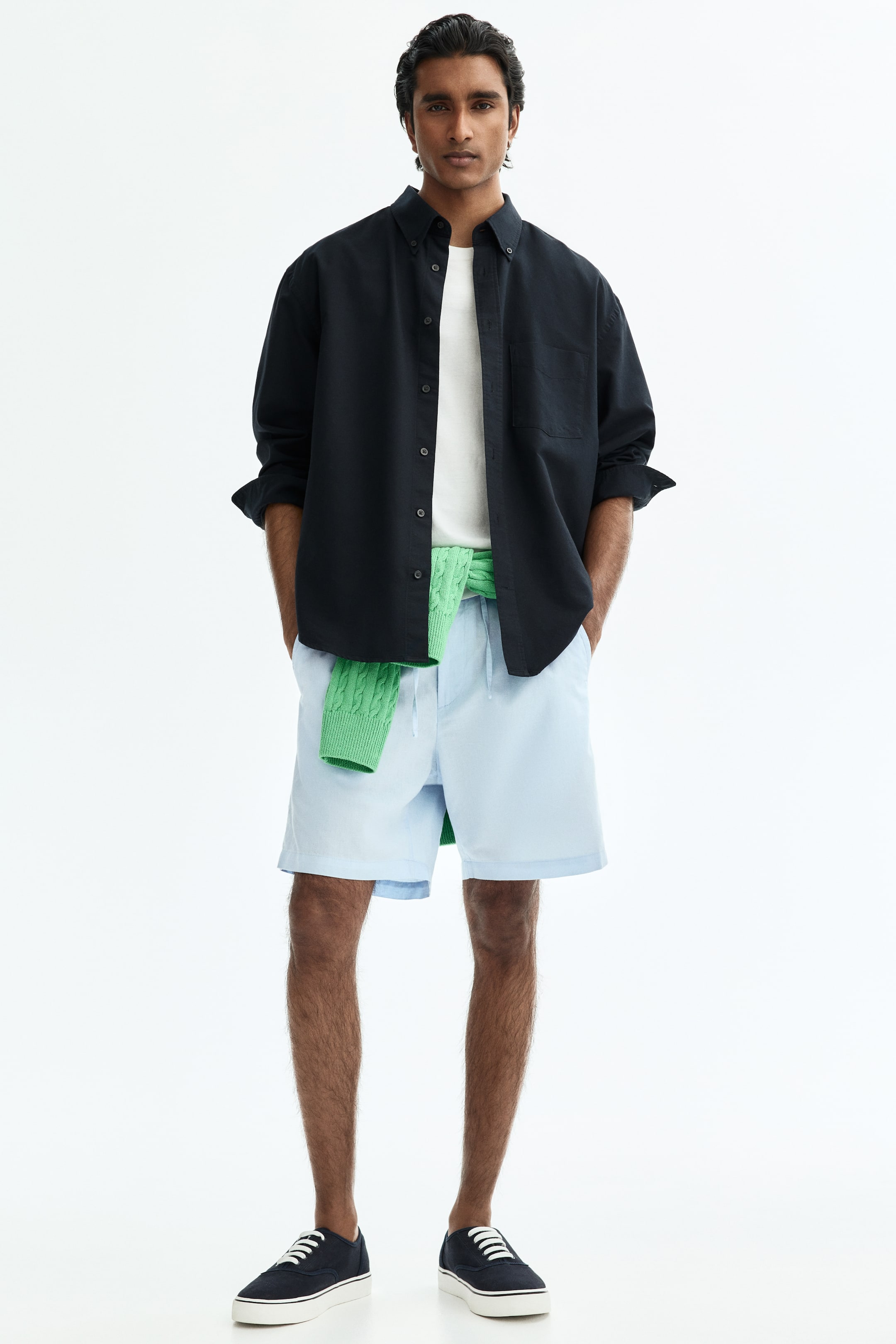 이미지 확대 보기: 검정색 버튼업 셔츠는 버튼다운 칼라, 단일 가슴 포켓, 팔뚝까지 말아 올린 긴 소매가 특징입니다. 흰색 티셔츠 위에 오픈 스타일로 착용하고 있습니다.