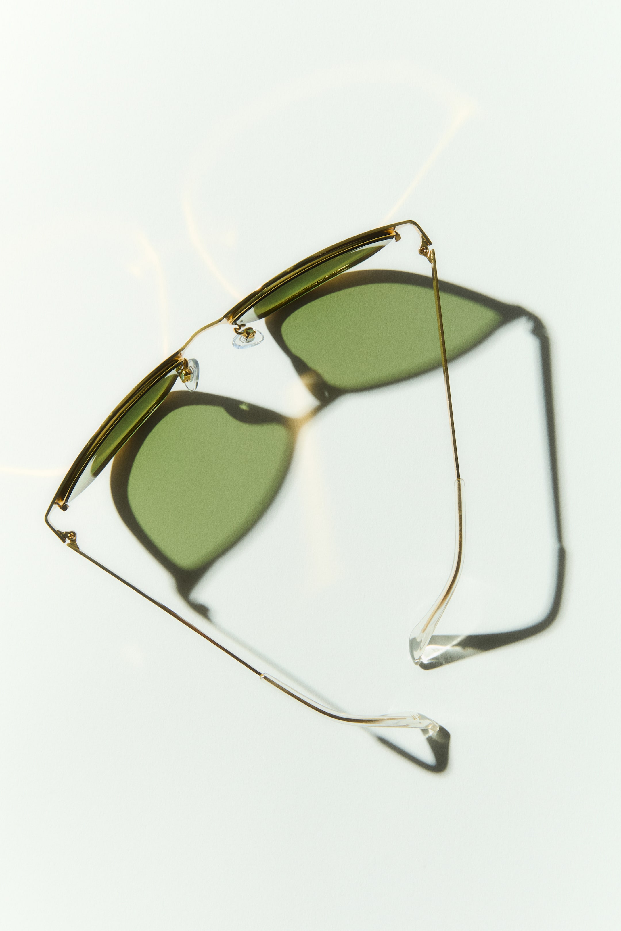 View larger image: The sunglasses boast a gold-toned metal frame with green lenses, set at an angle. Clear nose pads are visible, and a slender gold-toned temple with a clear ear tip extends to the side.
