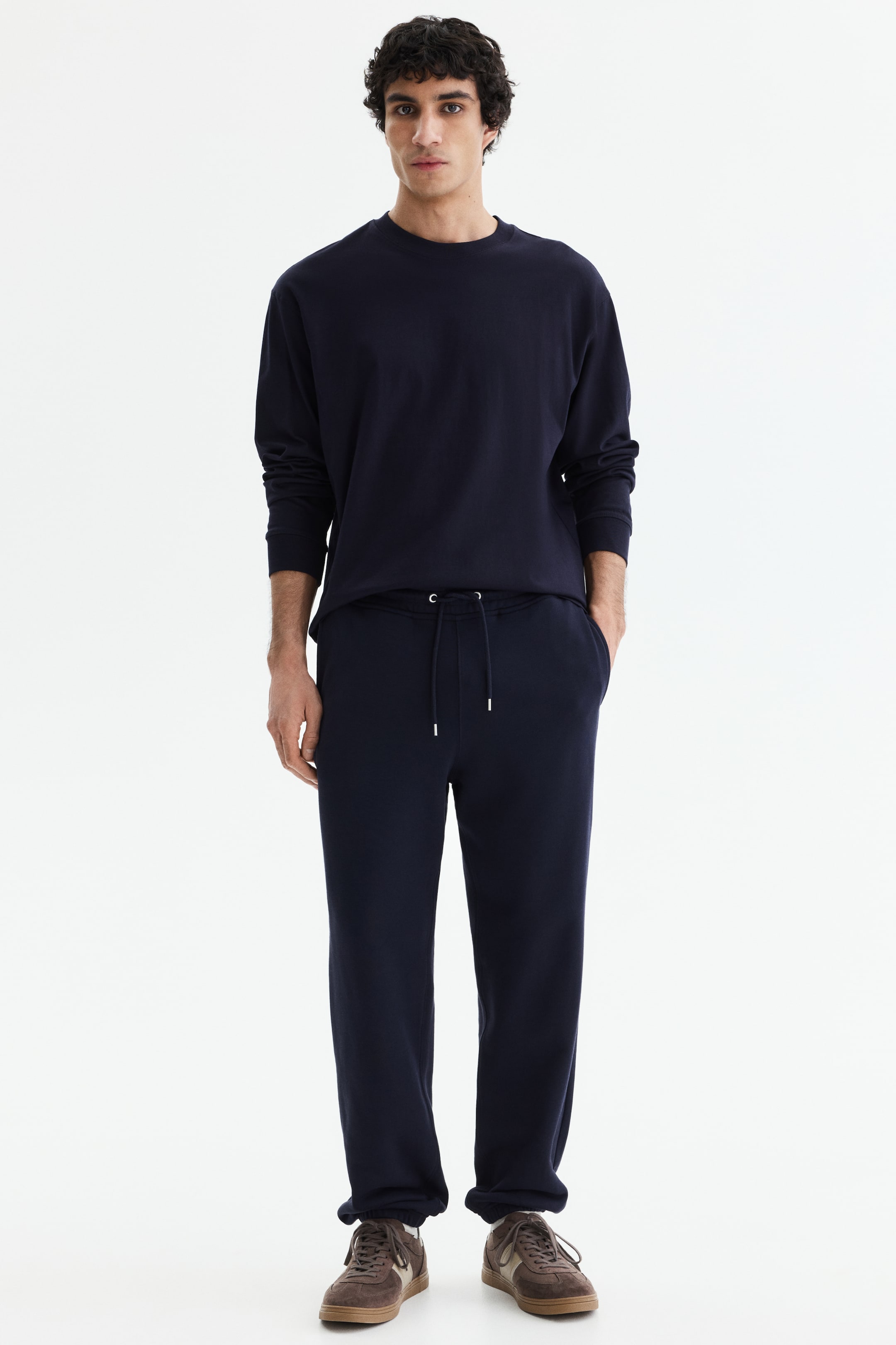 拡大画像を表示: ネイビーブルーの長袖クルーネックトップと、同じくネイビーブルーのスウェットパンツが正面向きに展示されています。スウェットパンツは、白いアグレット付きのドローストリングウエストバンド、伸縮性のある足首のカフスを特徴としており、片手がサイドポケットに入っています。