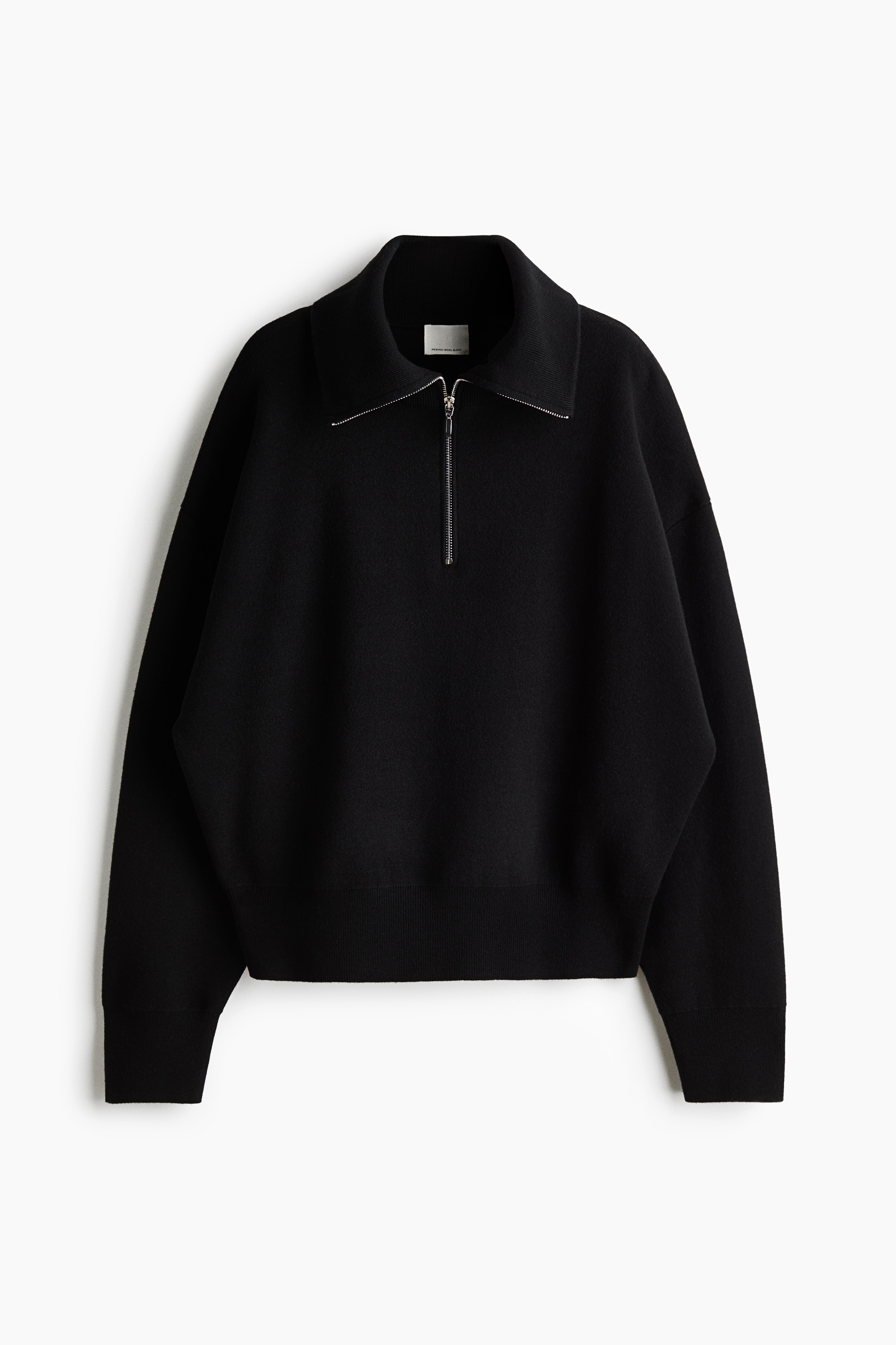 A black, fine-knit sweater featuring a ribbed funnel collar with a partially open silver half-zip is being worn by the model. The sweater is styled layered under a long, heavy black coat and over a white t-shirt, paired with matching black trousers.
