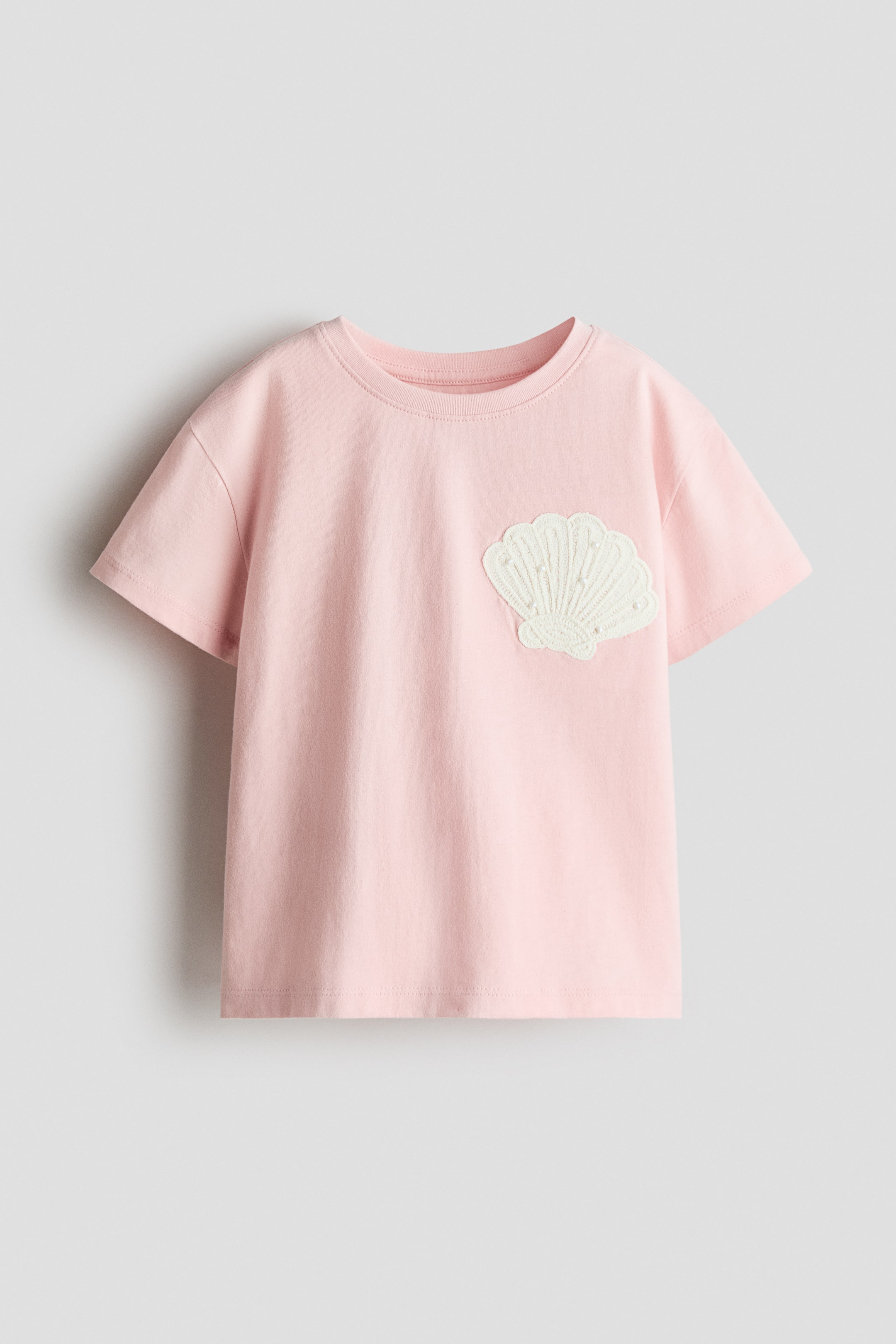 Agrandir l'image: Un t-shirt rose en jersey doux présente son devant, avec un col rond bordé de côtes, des épaules légèrement tombantes et un ourlet droit. Un appliqué de coquillage brodé de couleur crème avec de petits détails de perles orne le côté droit de l'avant.