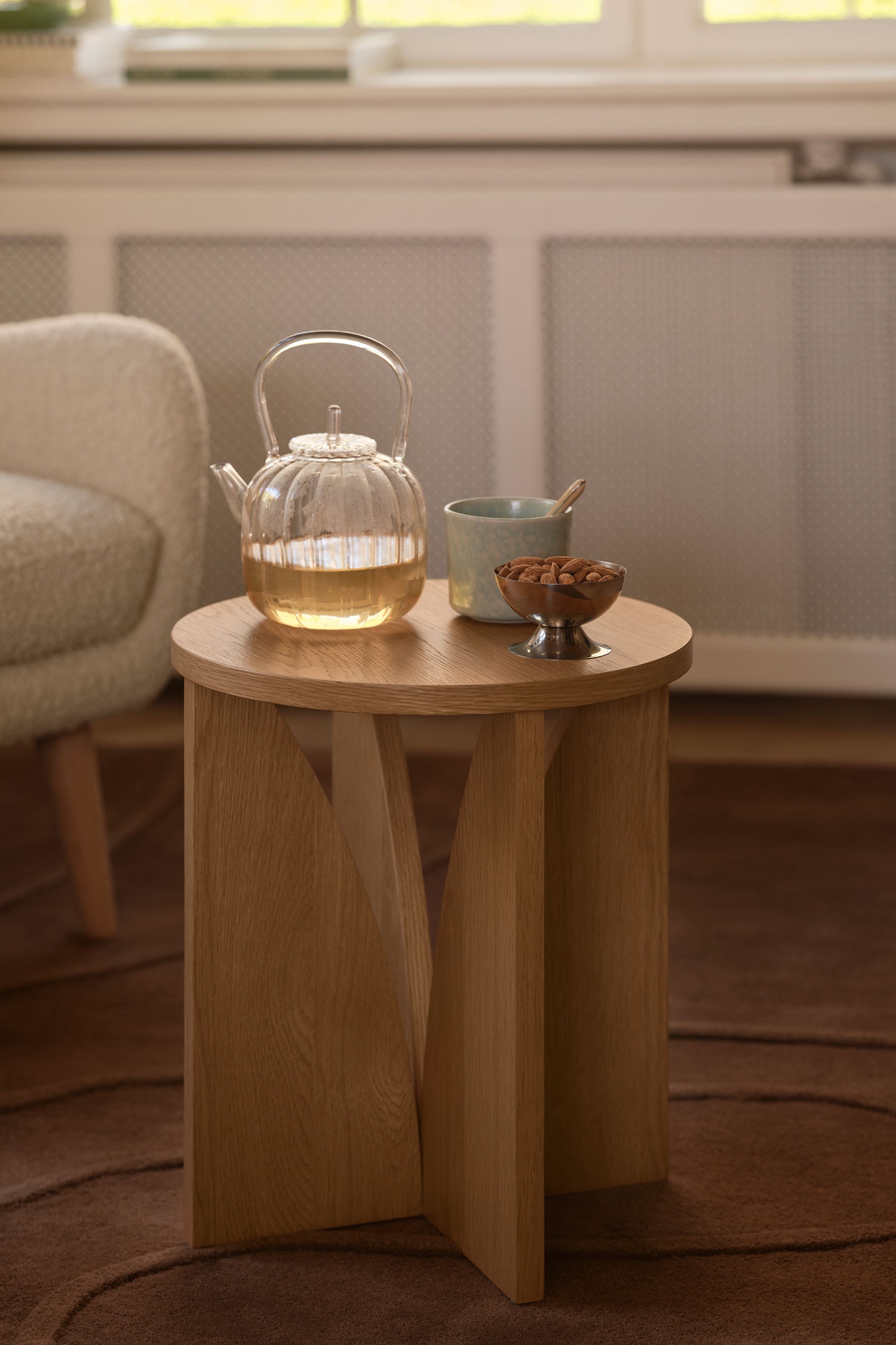 Een lichte eiken bijzettafel staat rechtop en heeft een vlak, rond tafelblad. De tafel wordt ondersteund door drie sculpturale, vlakke houten poten die naar binnen buigen en samenkomen aan de basis, waardoor een opvallend geometrisch silhouet ontstaat.