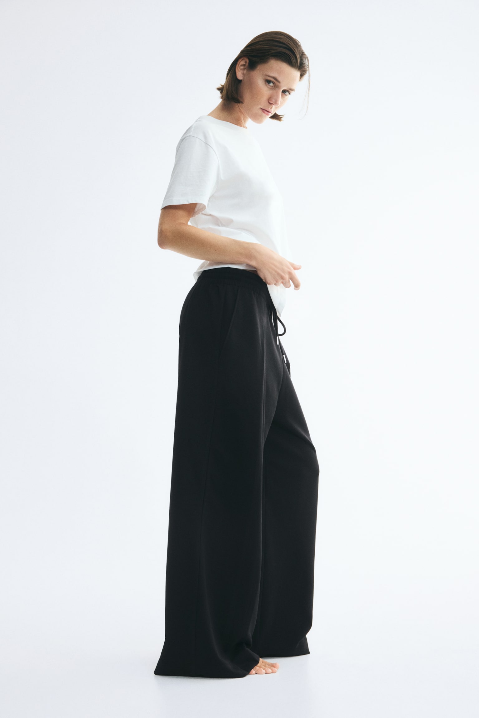 The black trousers feature an elasticated, gathered waistband with visible stitching. A braided drawstring emerges from the waist, crafted from a lightweight sweatshirt fabric. - 1