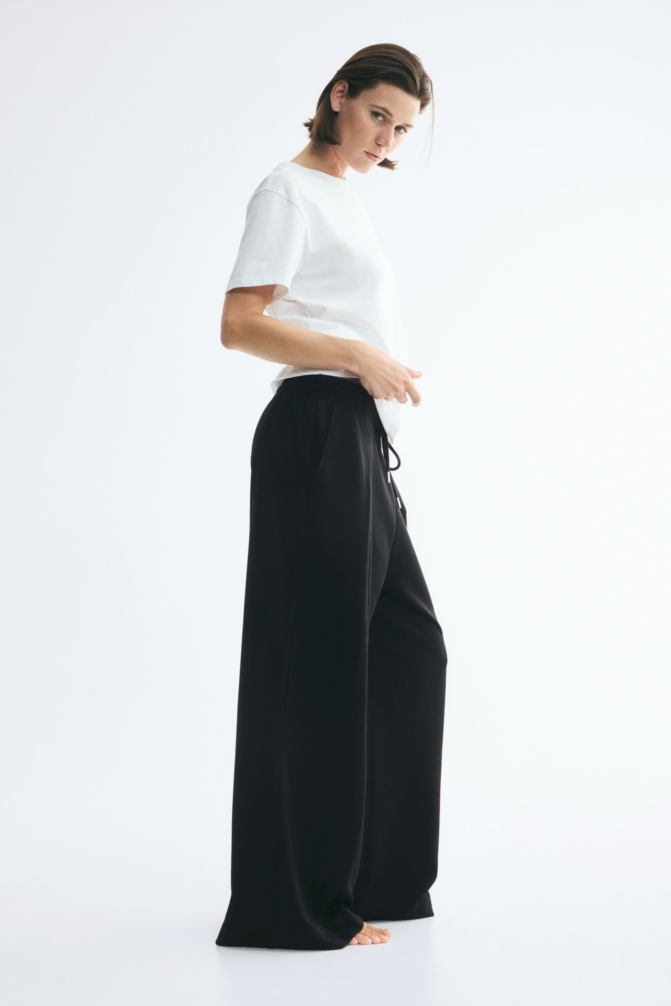 Agrandir l'image: Le mannequin porte un pantalon de jogging noir à coupe ample, doté d’une taille élastiquée avec cordon de serrage et de jambes extra-longues et larges avec pli sur le devant. Le mannequin associe ce pantalon à un t-shirt blanc à manches courtes, en se tenant de profil.