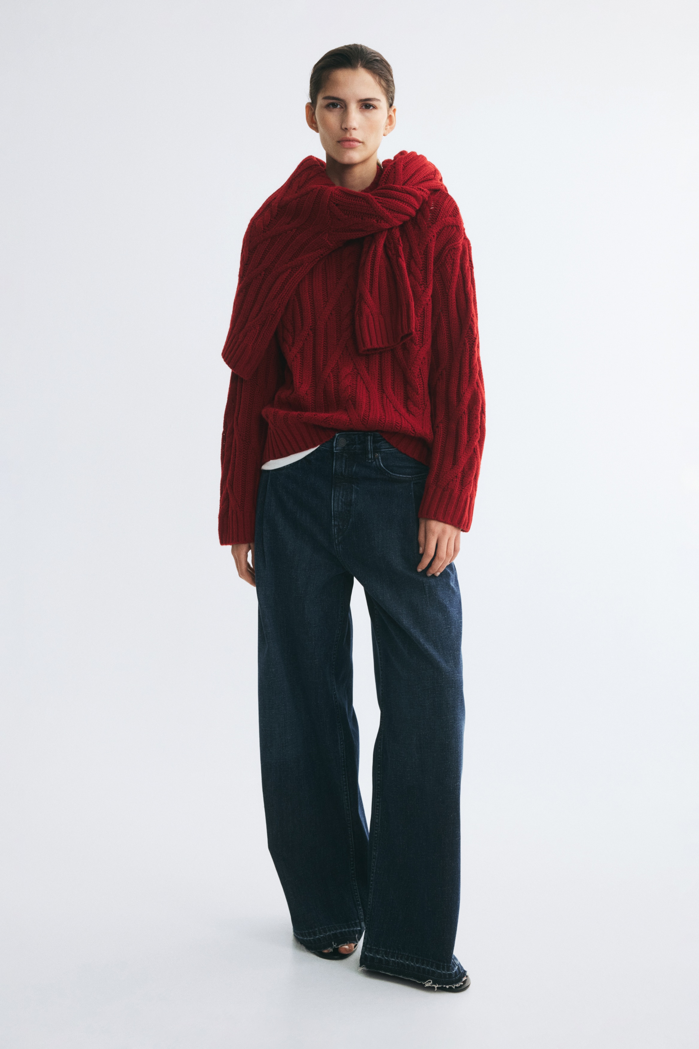 A red cable-knit sweater is shown from the front, displaying a thick diamond and twisted cable pattern across the body and long sleeves. The sweater features a ribbed mock neck, ribbed cuffs, and a ribbed hem.