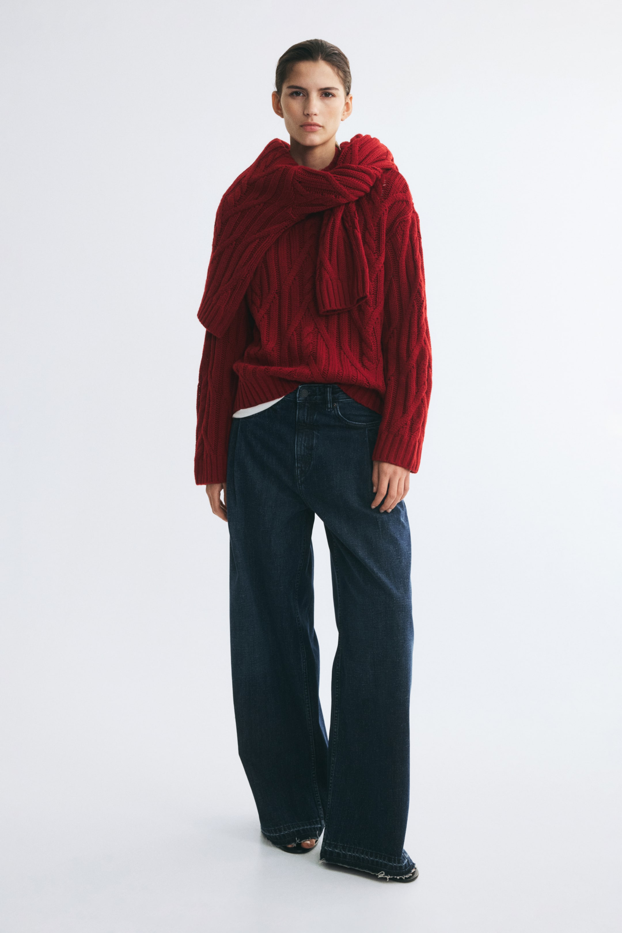 View larger image: The model is wearing a red oversized cable-knit sweater with a ribbed mock neck and dropped shoulders. A second matching red sweater is draped over the shoulders, styled with dark blue wide-leg jeans.