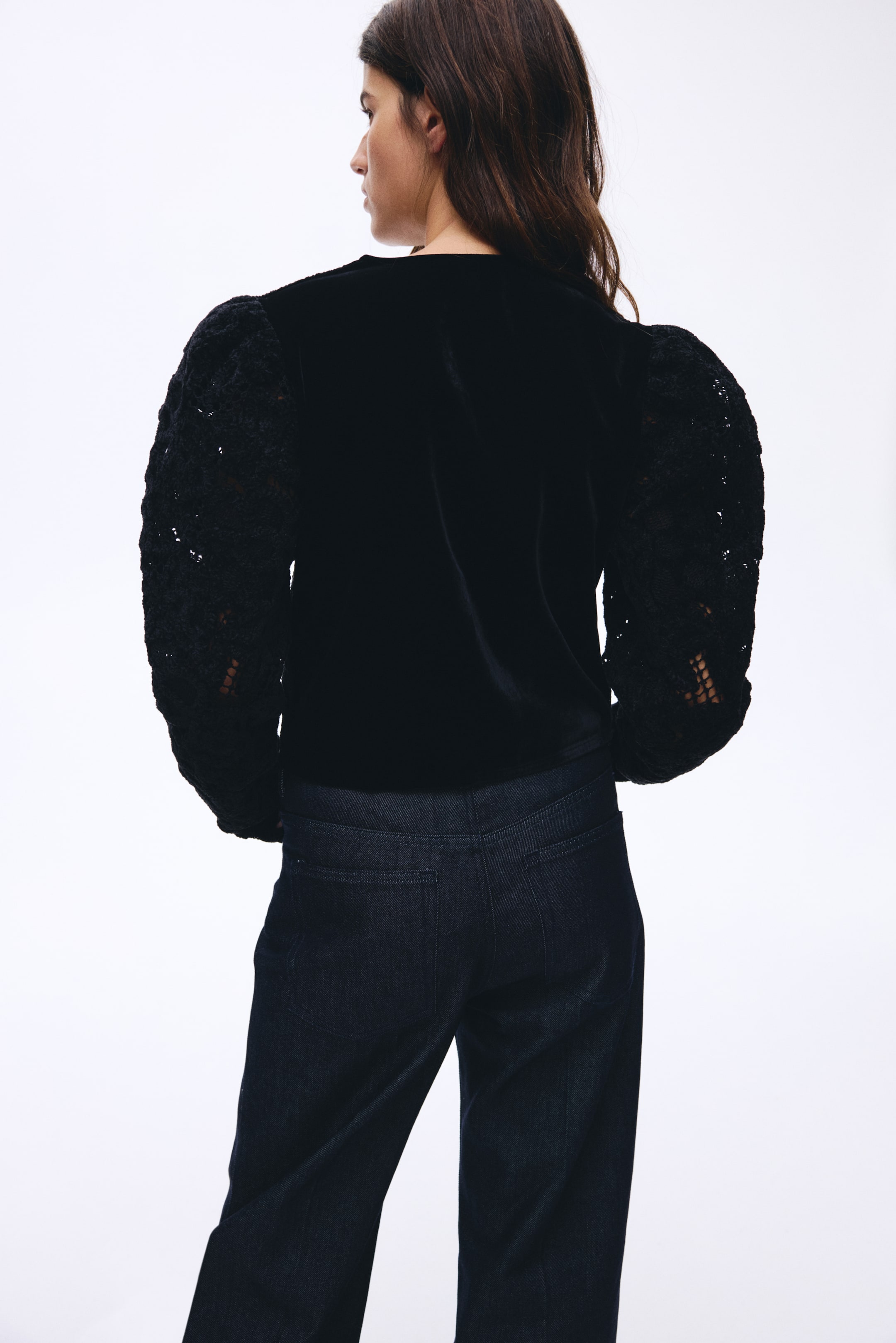 Größeres Bild ansehen: Das Produkt ist nach hinten gerichtet, leicht nach rechts geneigt und zeigt ein schwarzes Oberteil und dunkle Jeans. Das Oberteil besteht aus einem samtähnlichen Material, und seine voluminösen Puffärmel sind aus aufwendiger schwarzer Spitze gefertigt, während die dunklen Jeans einen geraden Schnitt mit sichtbaren Gesäßtaschen präsentieren.