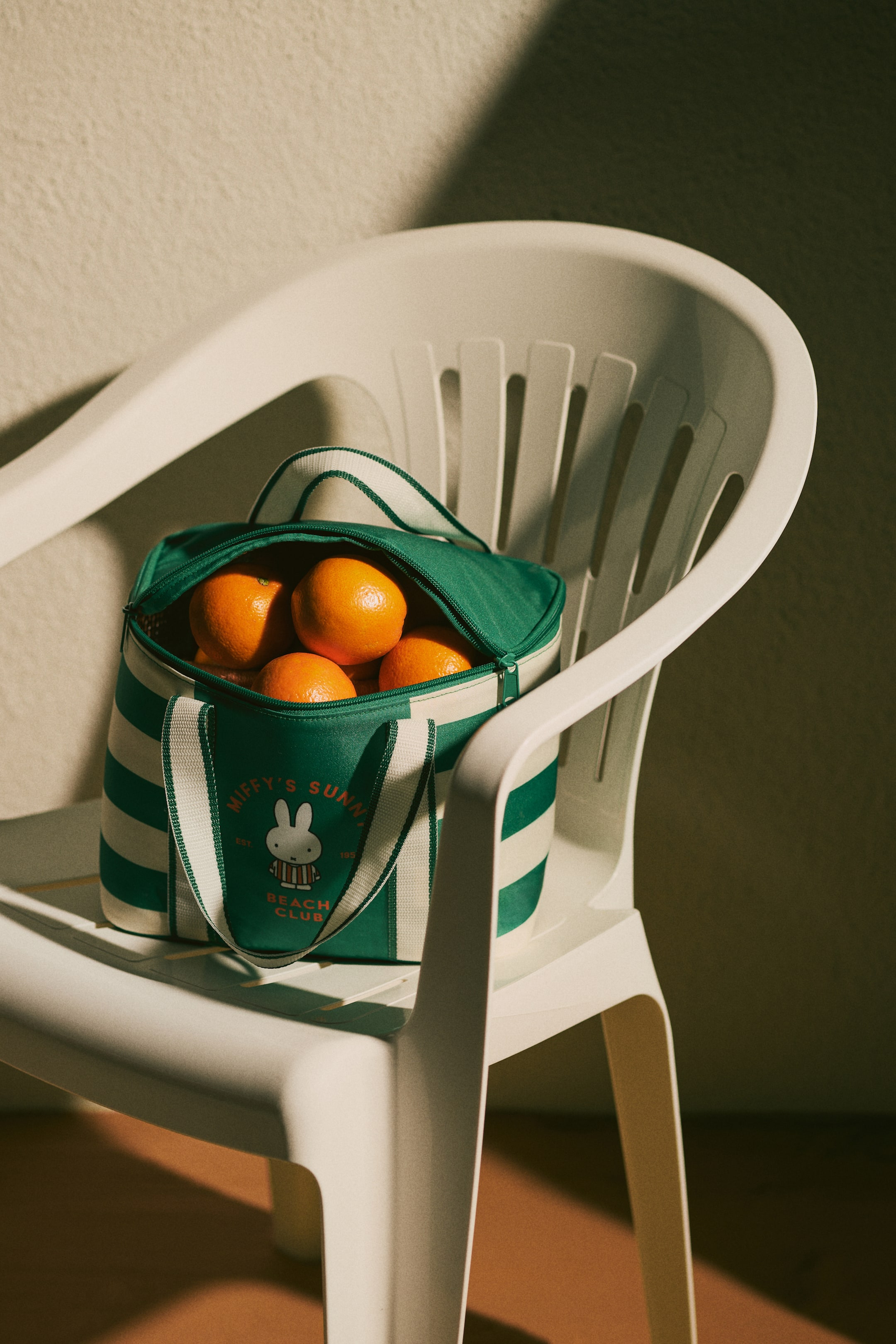Voir une image plus grande: Un sac isotherme à rayures vertes et blanches est positionné, sa fermeture éclair supérieure partiellement ouverte, révélant plusieurs oranges à l'intérieur. Le sac arbore un logo circulaire sur le devant, mettant en vedette un personnage de lapin blanc et le texte "Miffy's Sunny Beach Club."