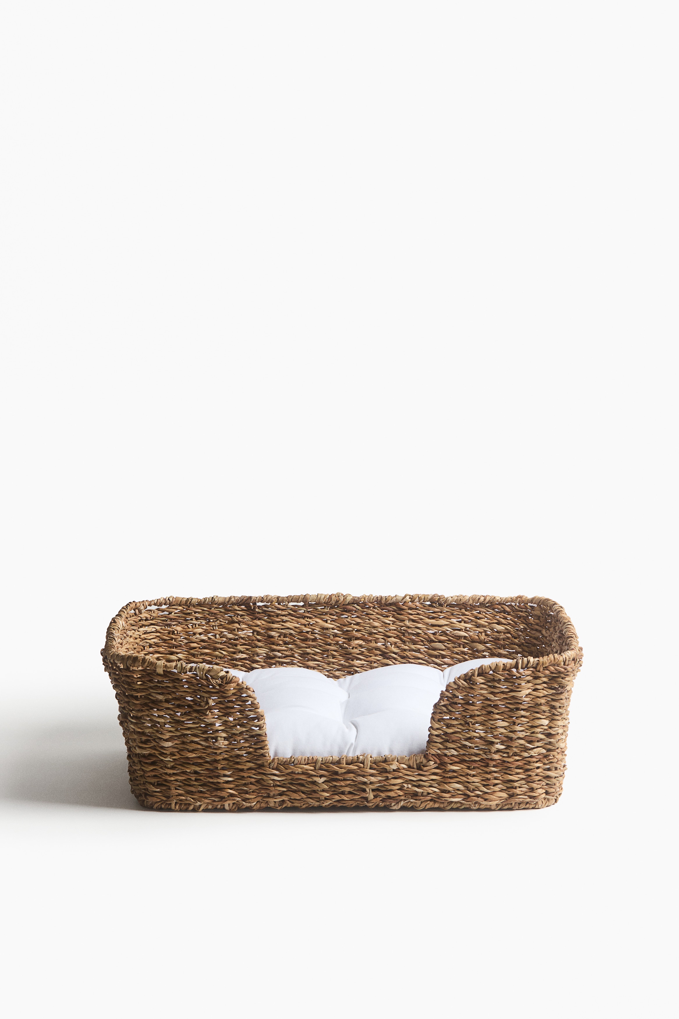 Un panier pour animal tissé est orienté de face, mettant en valeur une structure rectangulaire en fibres naturelles tissées avec une entrée frontale abaissée. Le panier contient un coussin moelleux blanc capitonné, parfaitement ajusté à l’intérieur.