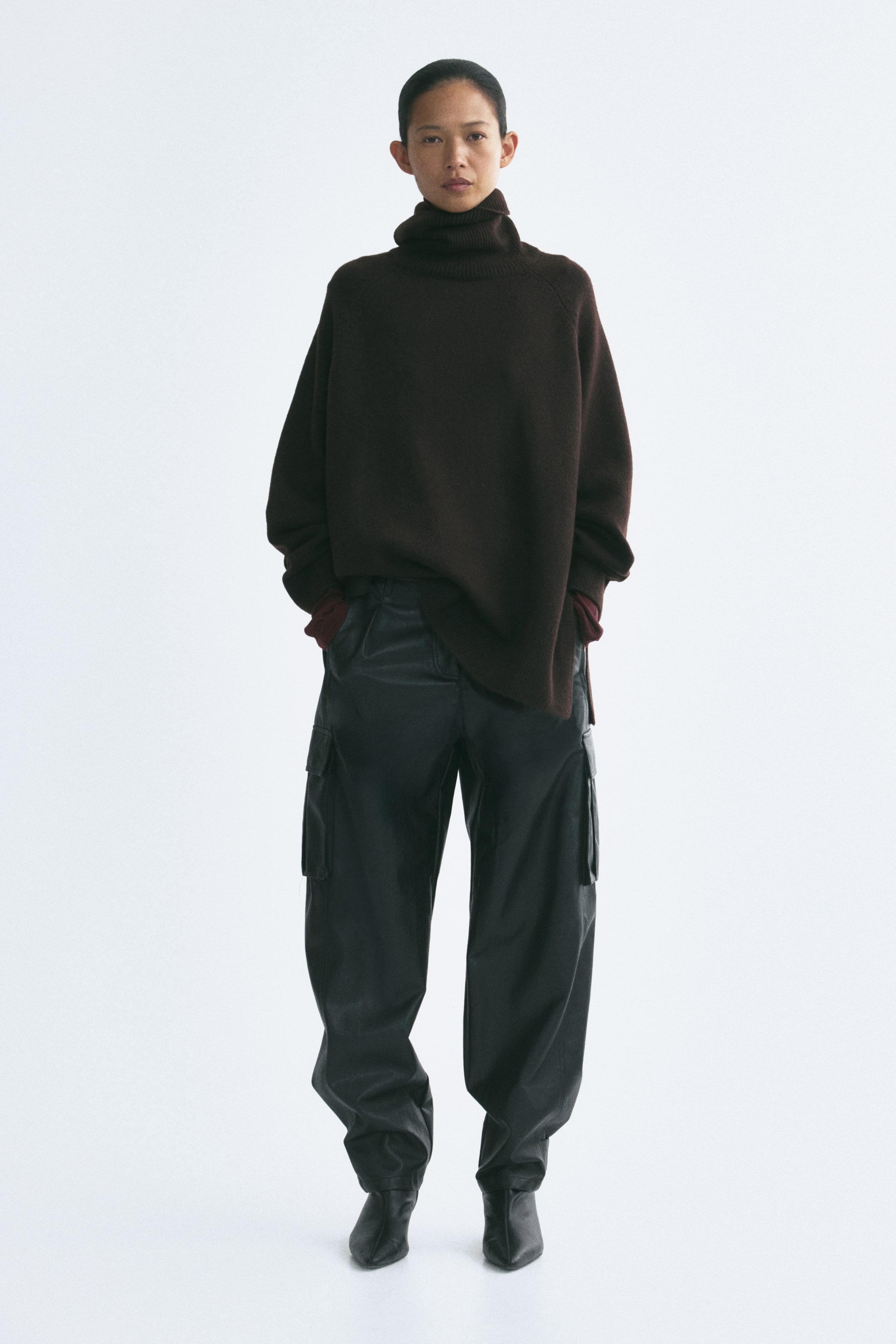 View larger image: An oversized dark brown knit jumper featuring a ribbed roll neck, long raglan sleeves with ribbed cuffs, and a ribbed hem with side slits is being worn by the model. The model is standing with their hands in the pockets of black cargo trousers, causing the hem of the jumper to drape over the waistband.