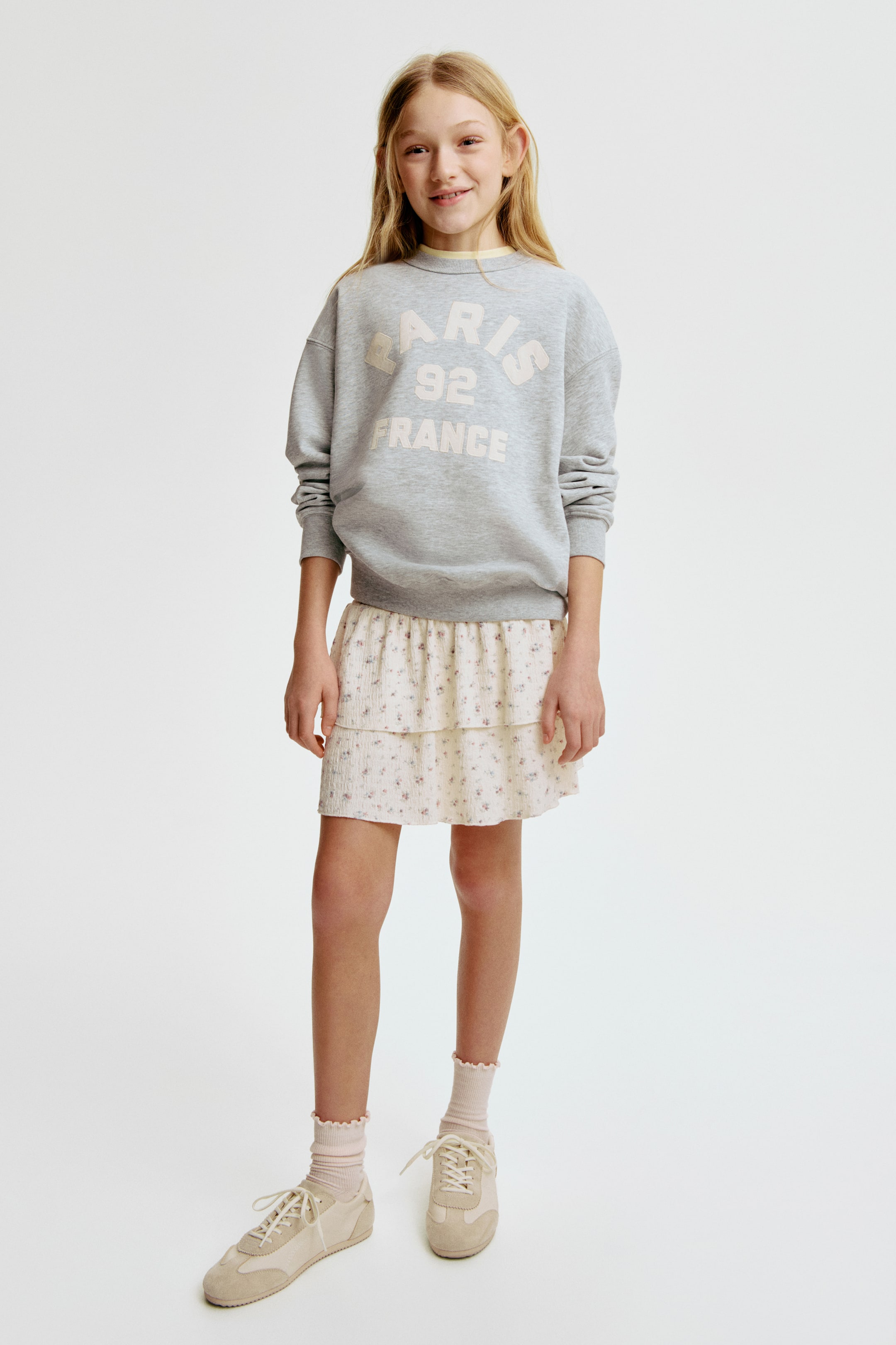 View larger image: A gray crew neck sweatshirt with ribbed cuffs and hem, displaying "PARIS 92 FRANCE" text in light beige, is worn over a visible yellow collar and a light cream tiered skirt featuring a small floral print. Light pink ruffled ankle socks and beige lace-up sneakers complete the outfit.