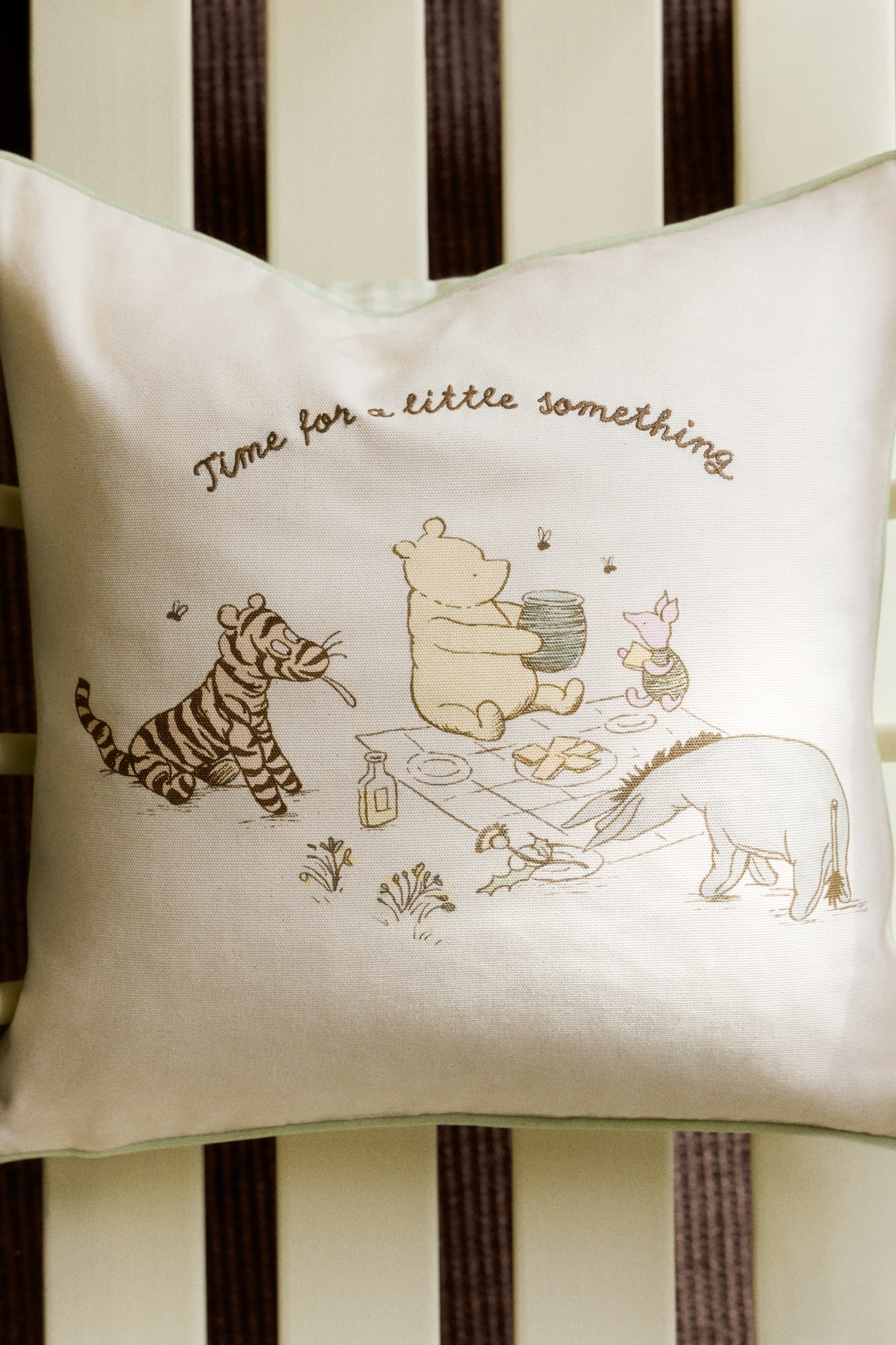 A cushion cover crafted from cotton canvas showcases a printed motif on the front, depicting Winnie the Pooh characters enjoying a picnic. Embroidered text reading "Time for a little something" elegantly arches above the scene, with accent-colour piping outlining the edges. - 1