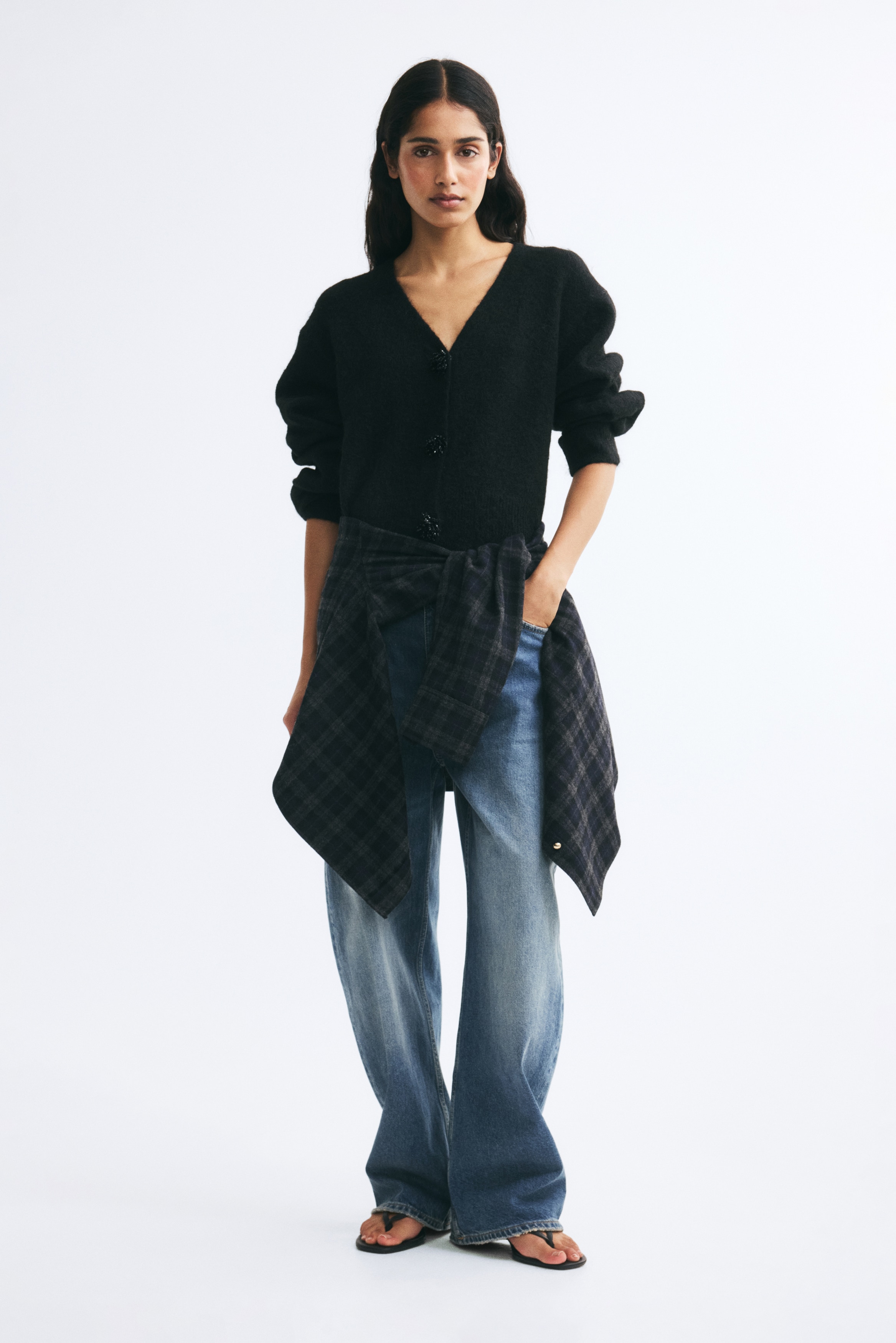 A black knit cardigan is shown from the front, featuring a fuzzy texture, a V-neckline, and long voluminous sleeves with ribbed cuffs. The cropped cardigan fastens down the centre with three large, embellished black buttons.