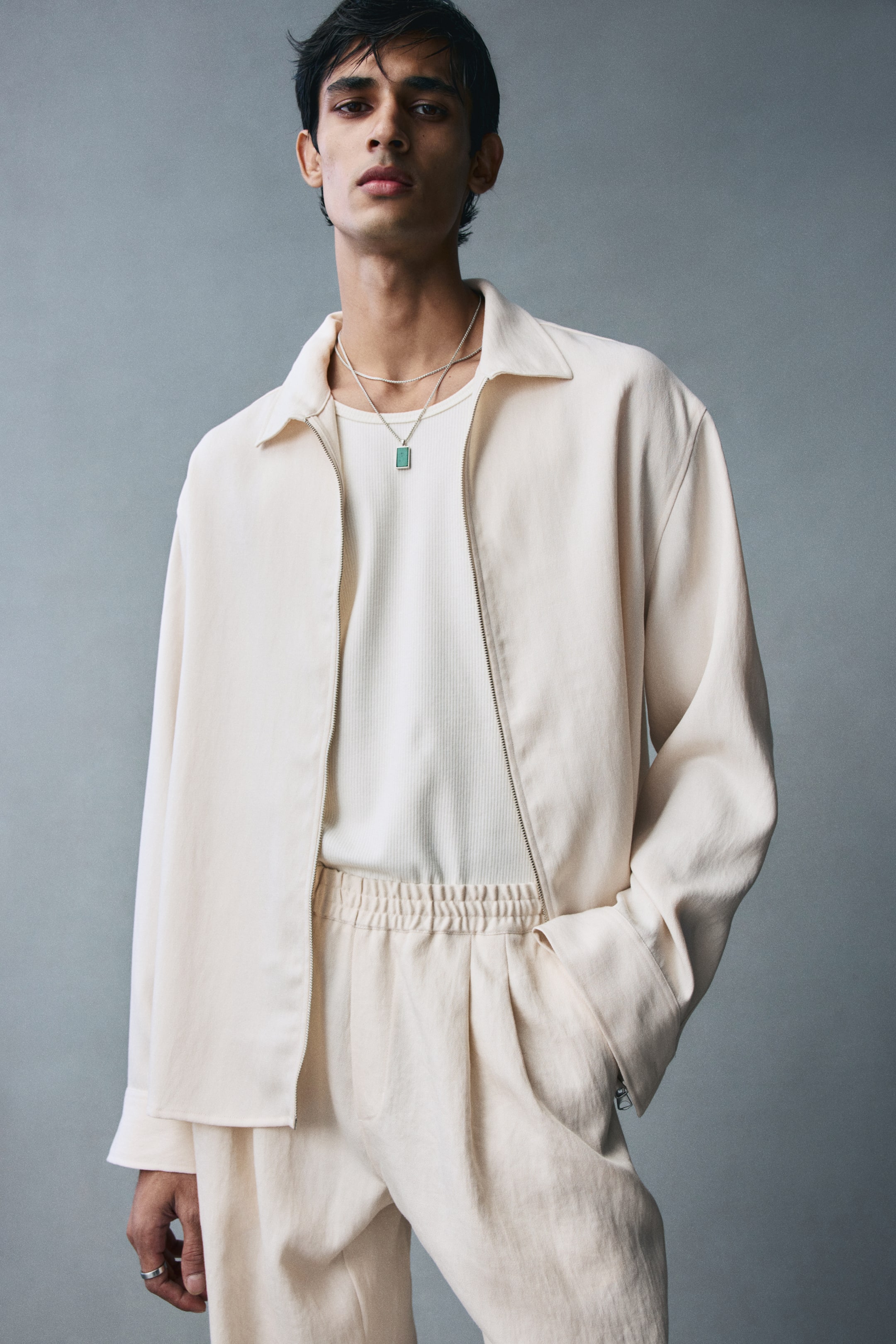 View larger image: Cream-coloured viscose-blend twill pants featuring a relaxed fit and a gathered elasticated waistband are worn by the model. The model is standing with one hand resting inside the diagonal side pocket of the pants, styled with a matching open jacket and a tucked-in ribbed tank top.