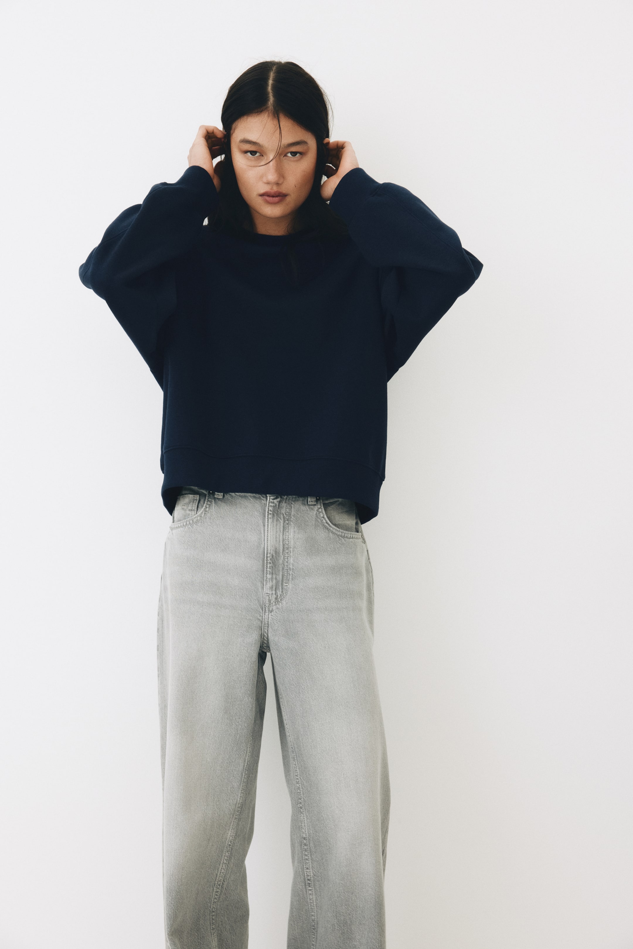 Nagyobb kép megtekintése: A modell egy túlméretezett sötétkék pulóvert visel, amely kerek, bordázott nyakkivágással, ejtett vállakkal és hosszú ujjakkal rendelkezik, bordázott mandzsettával és szegéllyel. A modell mindkét kezét a feje oldalához emeli, megérinti a haját, és világosszürke, bő szárú farmert is visel.