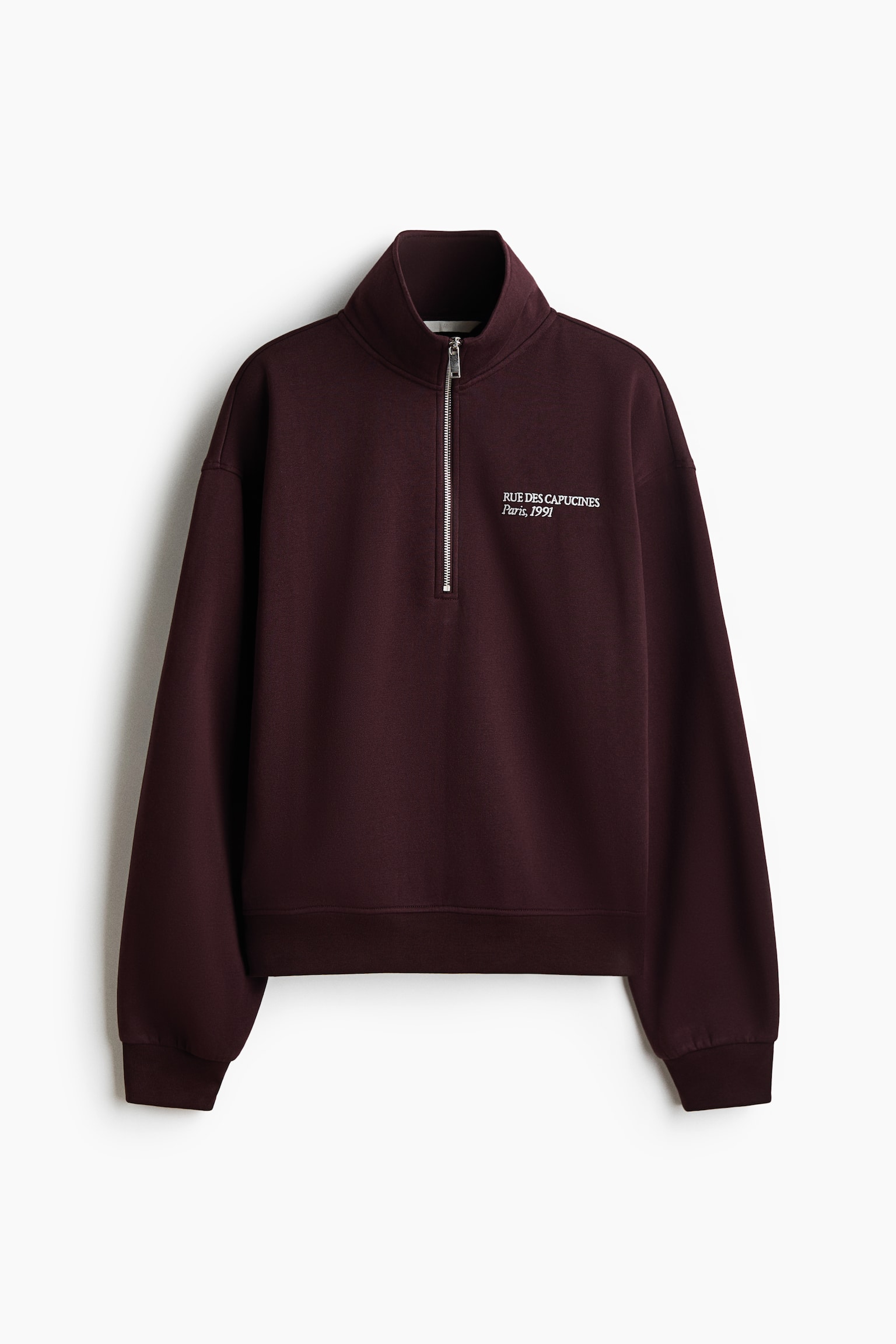 A dark burgundy fleece sweatshirt is shown from the front, featuring a stand collar with a silver-tone quarter-zip, dropped shoulders, and ribbed cuffs and hem. The sweatshirt displays small white printed text on the left chest that reads "RUE DES CAPUCINES Paris, 1991".
